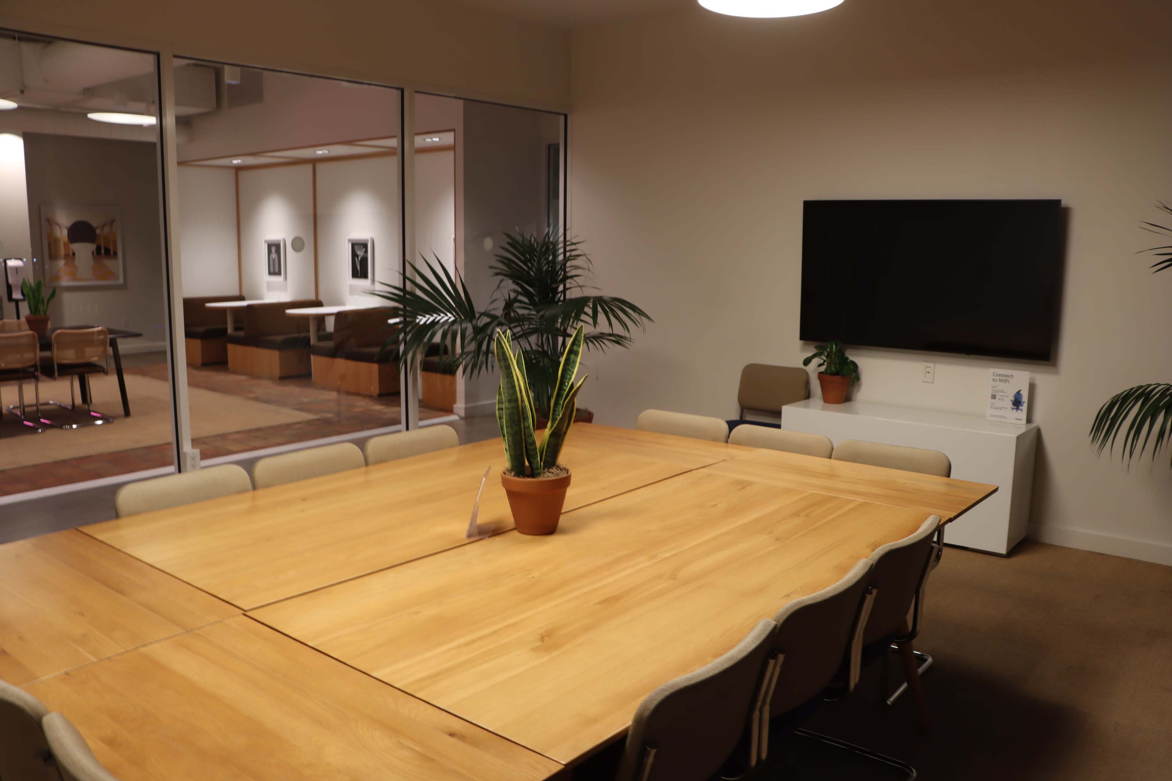 The image shows a large wooden conference table with chairs around it, a potted plant in the center, and a TV mounted on the wall in a well-lit meeting room.