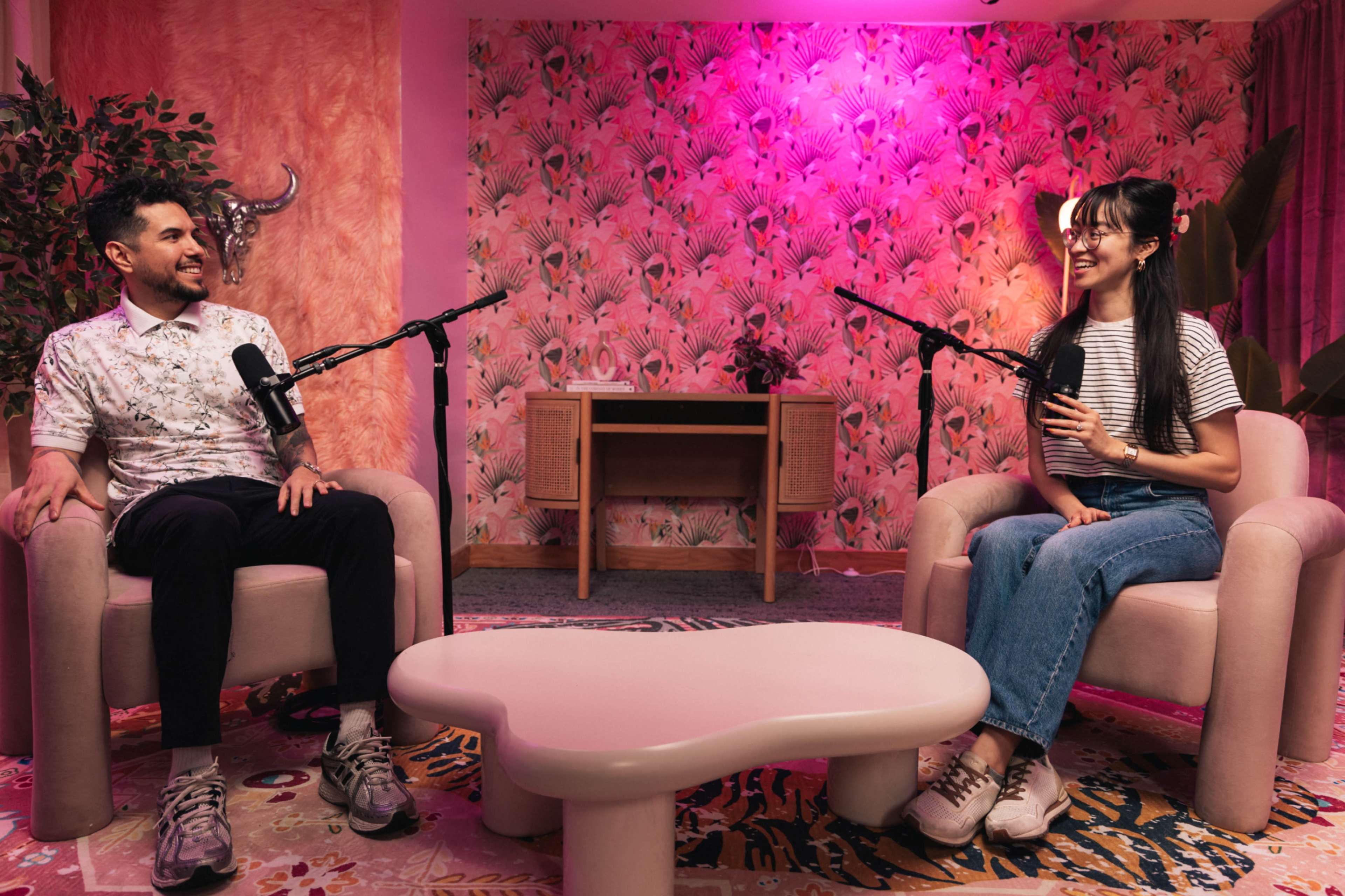 Two people sit in armchairs with microphones in a brightly lit, stylish room featuring floral wallpaper and a decorative table.