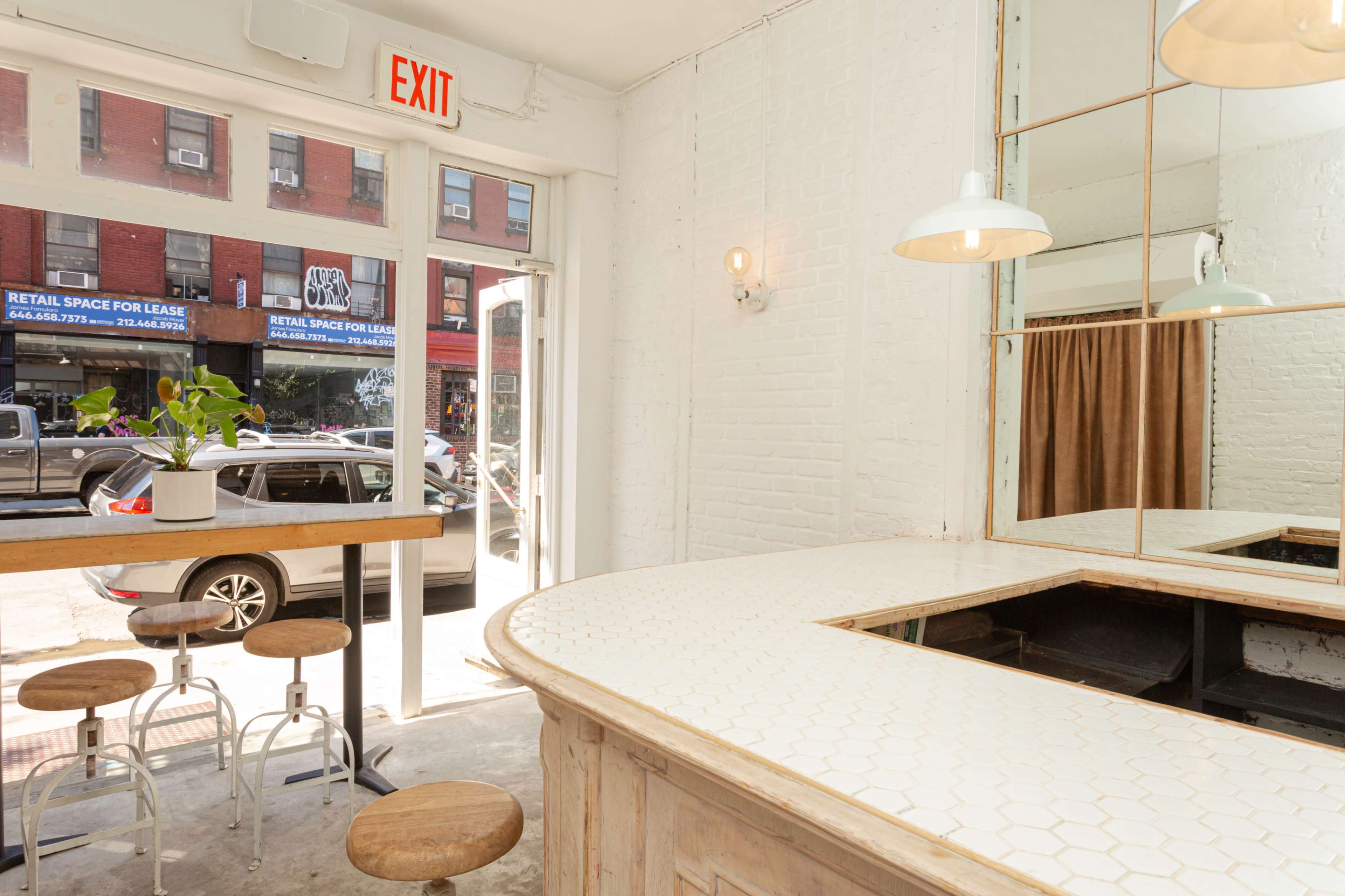 The image shows a bright interior of a cafe with a curved counter, wooden bar stools, and large windows overlooking a street with shops.