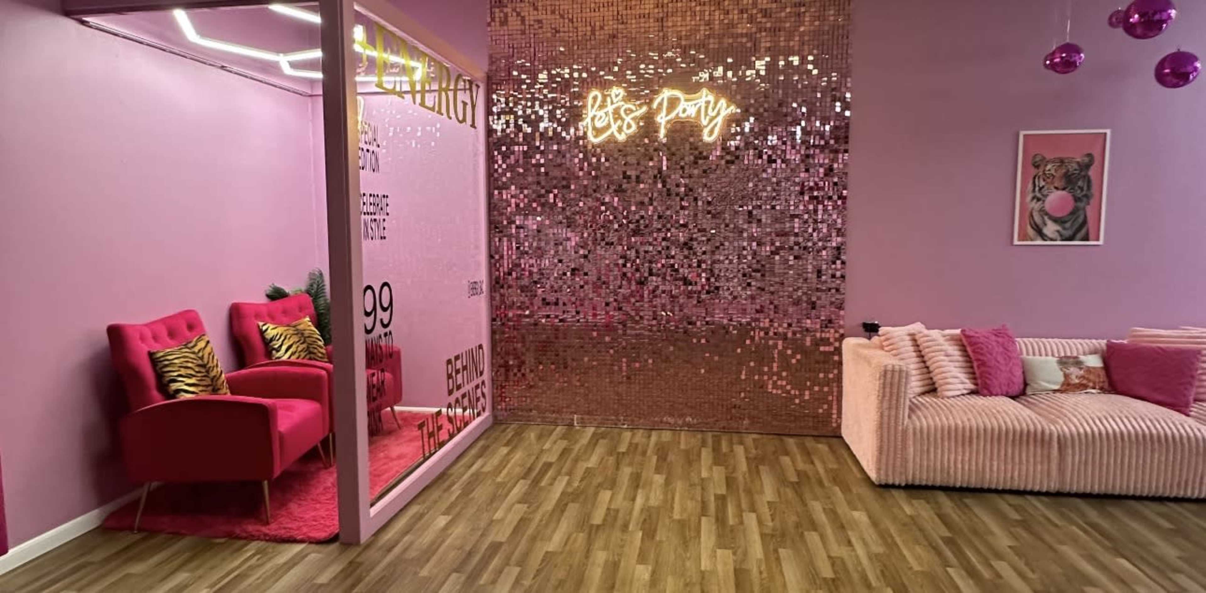 The image depicts a vibrant, pink-themed room featuring a seating area with two pink chairs and a wall covered in shiny, gold sequins.