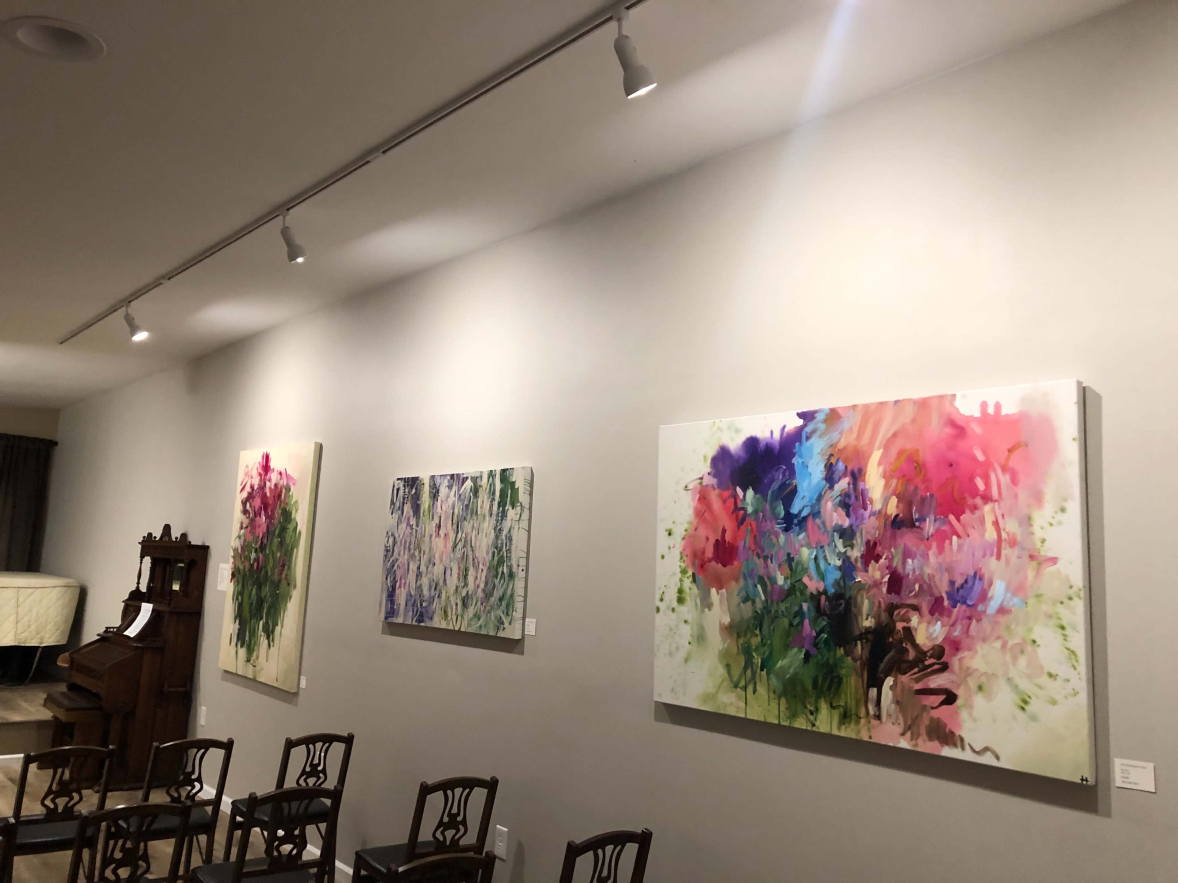 A gallery wall displaying three colorful abstract paintings, with wooden chairs arranged in front.