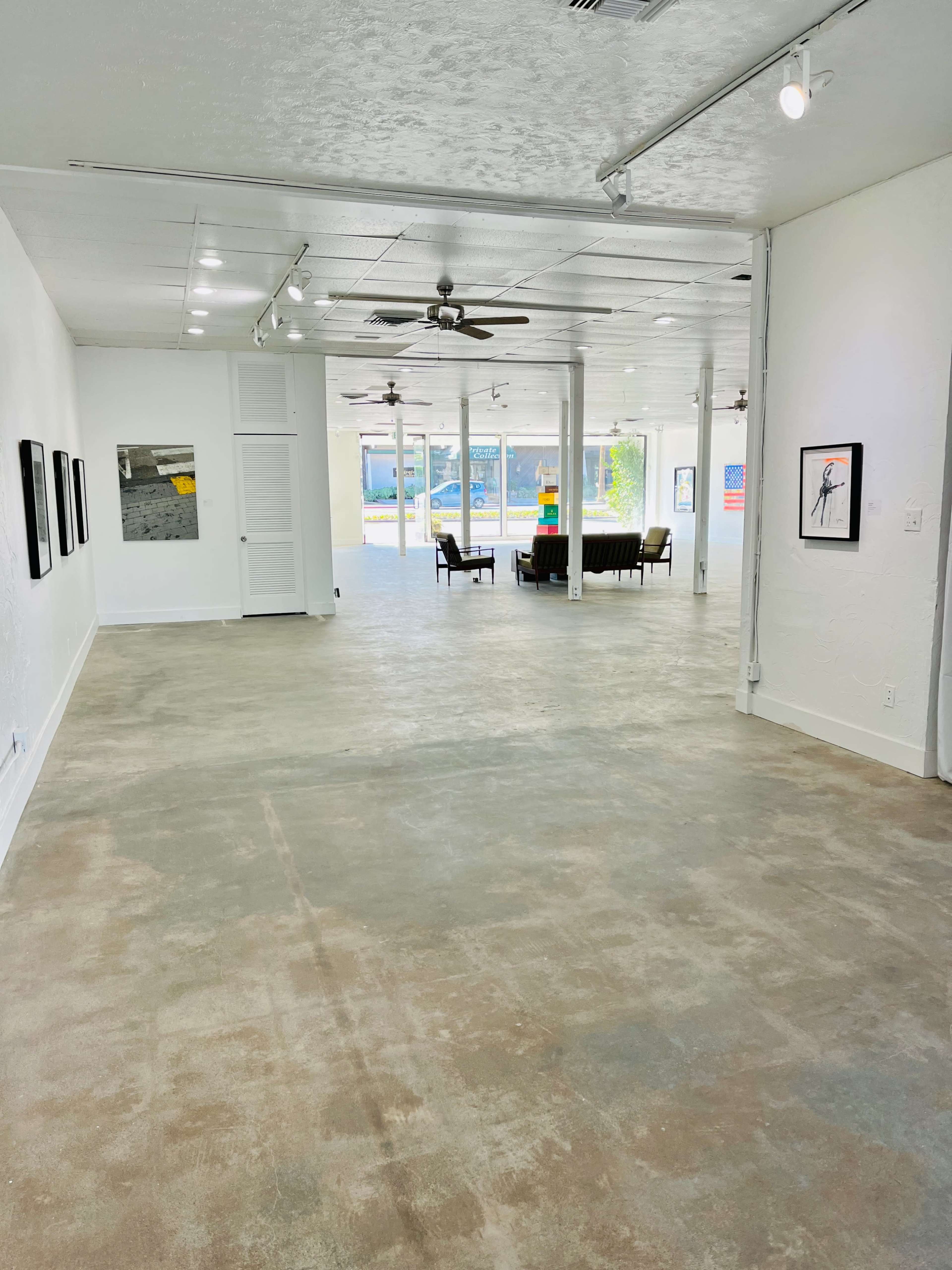 A spacious gallery with polished concrete flooring and large windows letting in natural light, featuring a few framed artworks on the white walls.
