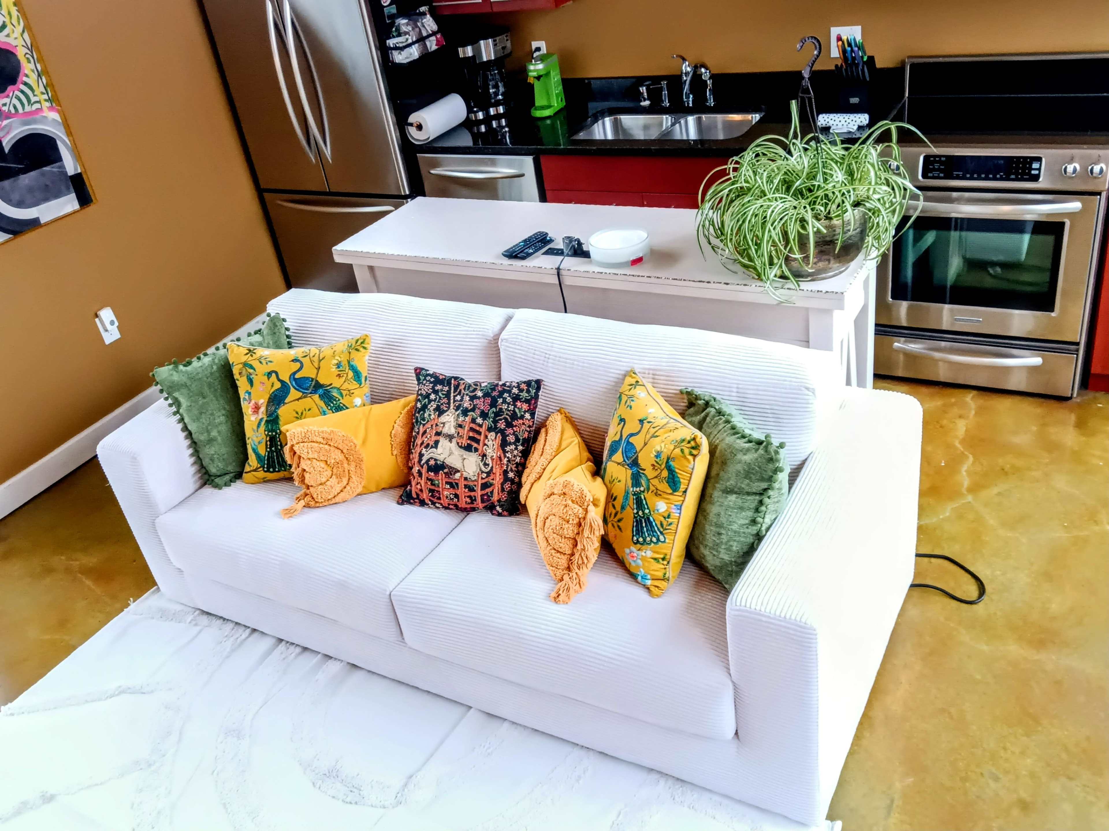 A white sofa adorned with colorful cushions, positioned in a kitchen area with stainless steel appliances and a countertop.