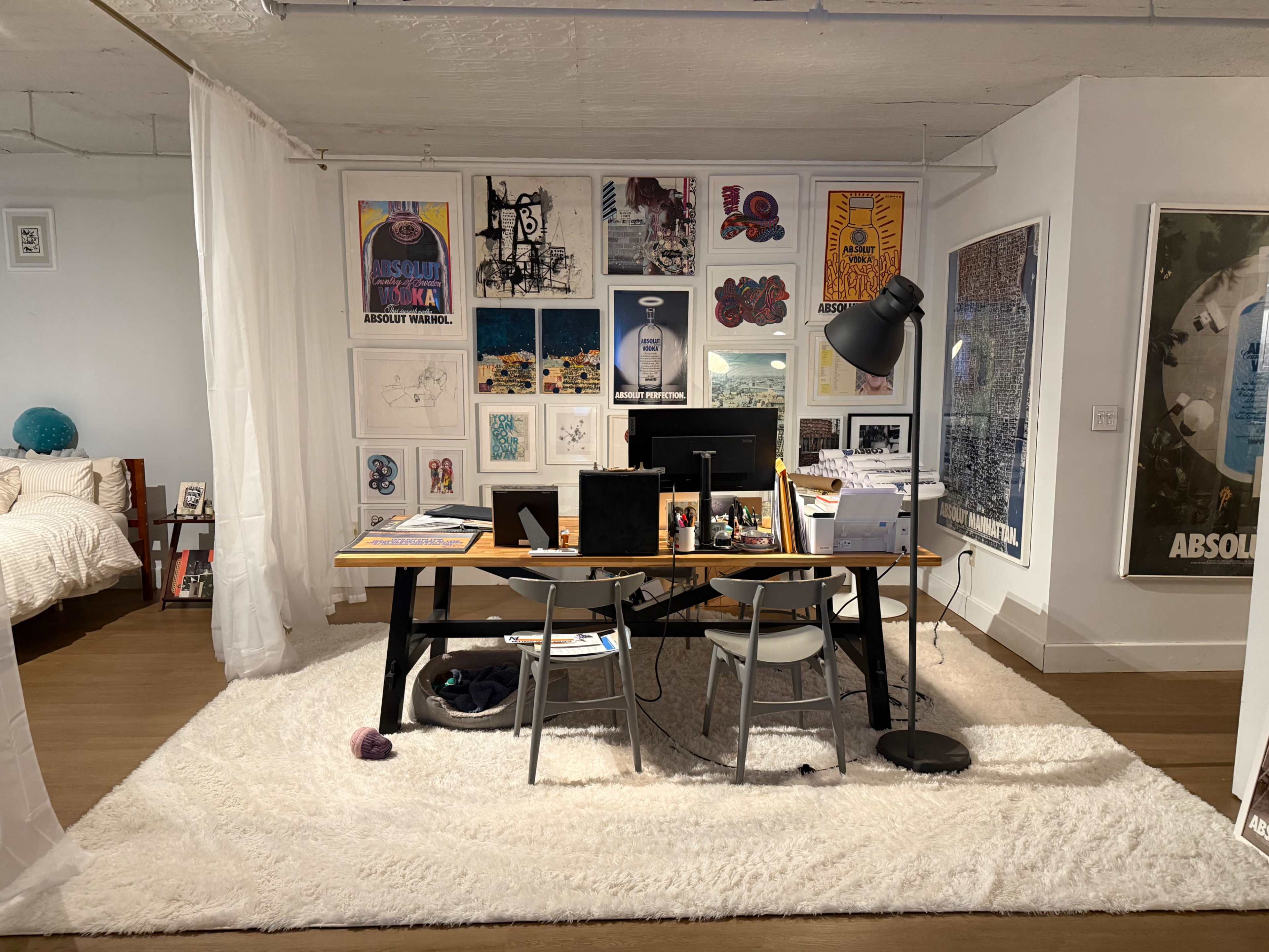 The image shows a modern home office space featuring a desk with a computer and art supplies, surrounded by framed artwork on the walls, and a bed partially visible in the background.