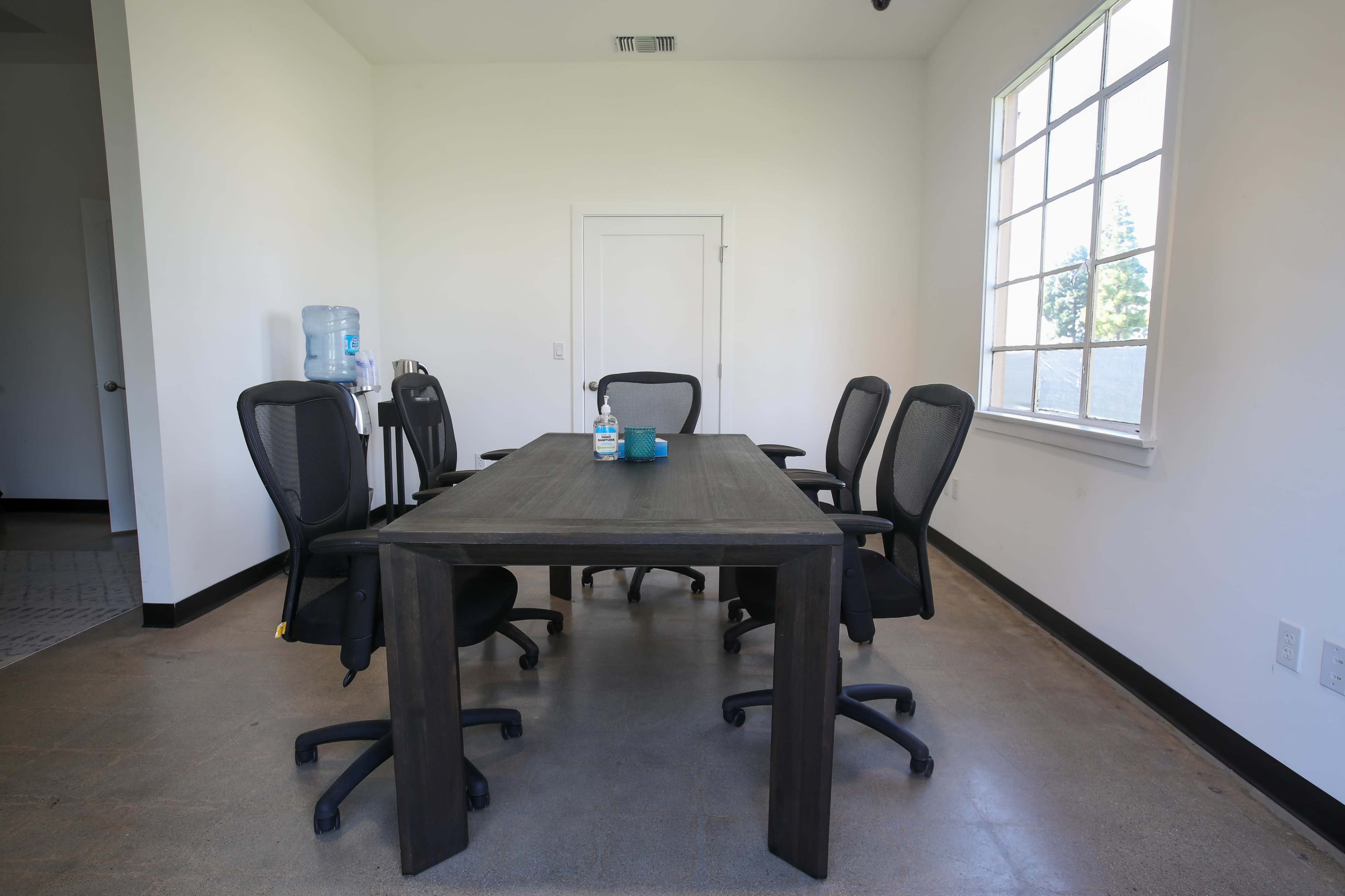 A conference room with a large wooden table surrounded by black office chairs, a water dispenser in the corner, and a window letting in natural light.
