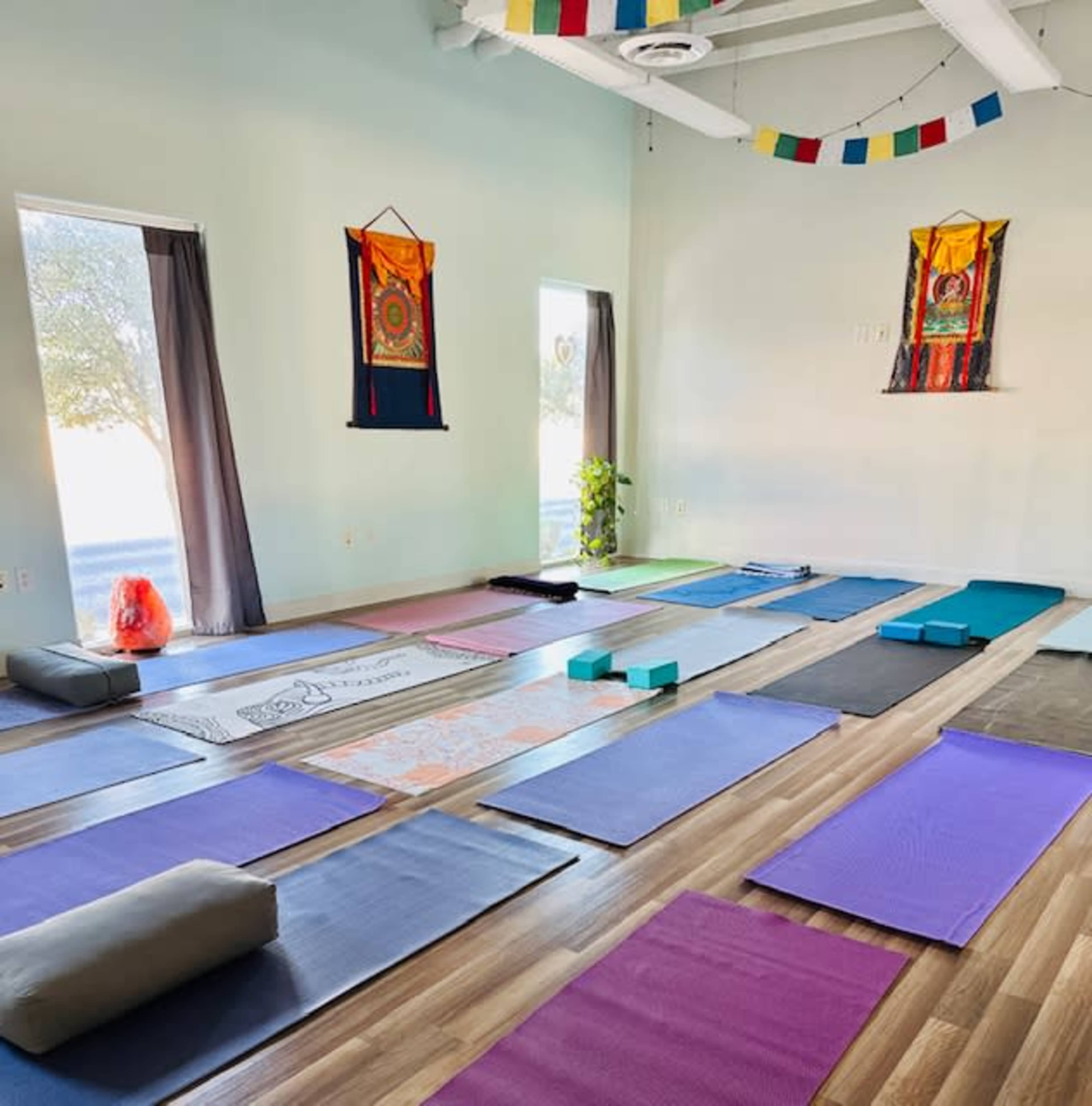 The image shows a yoga studio with colorful mats arranged in rows, surrounded by decorative walls and natural light coming through windows.