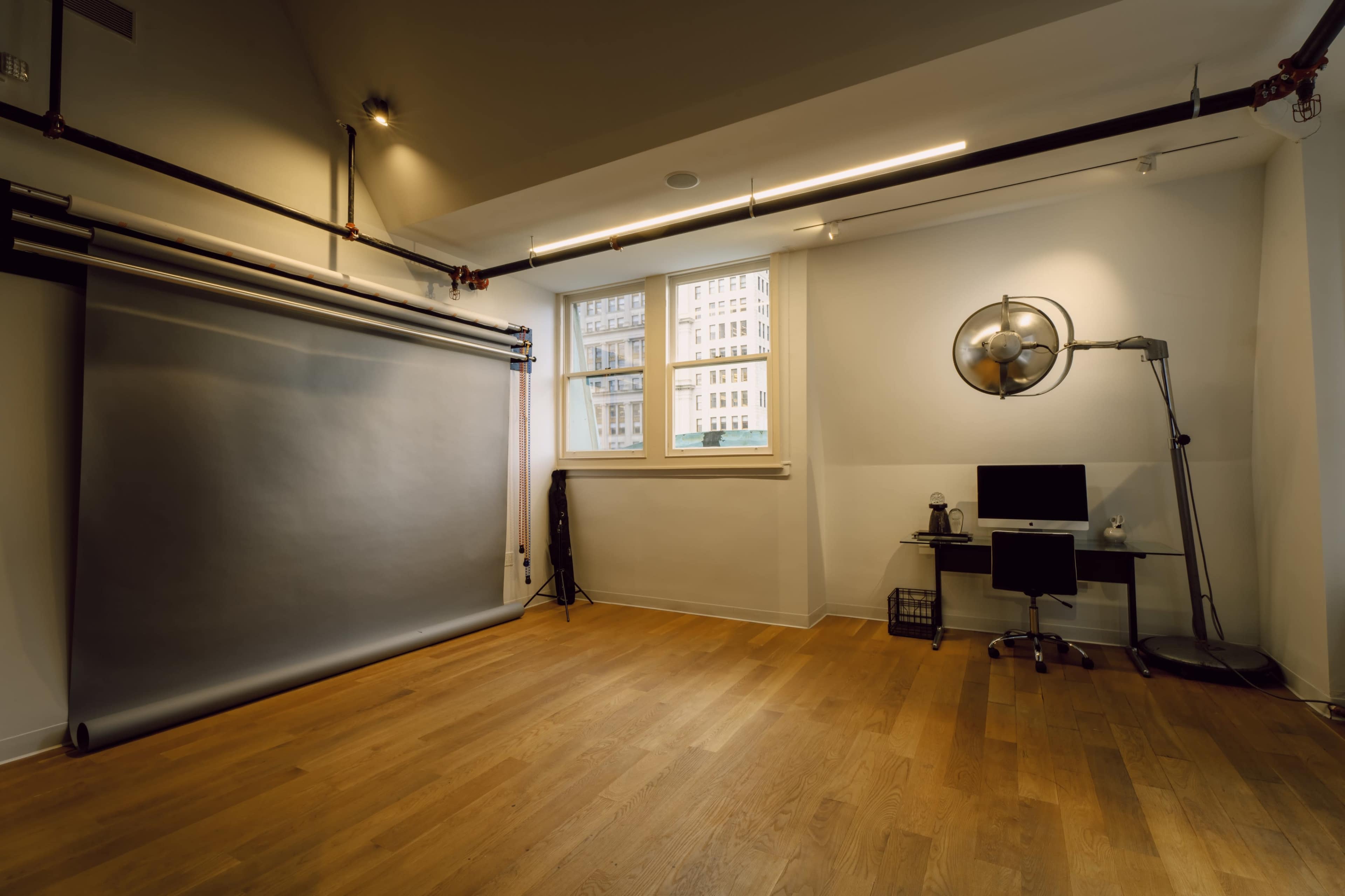 The image depicts a minimalist studio space featuring a grey backdrop, a small desk with a computer, and large windows allowing natural light.