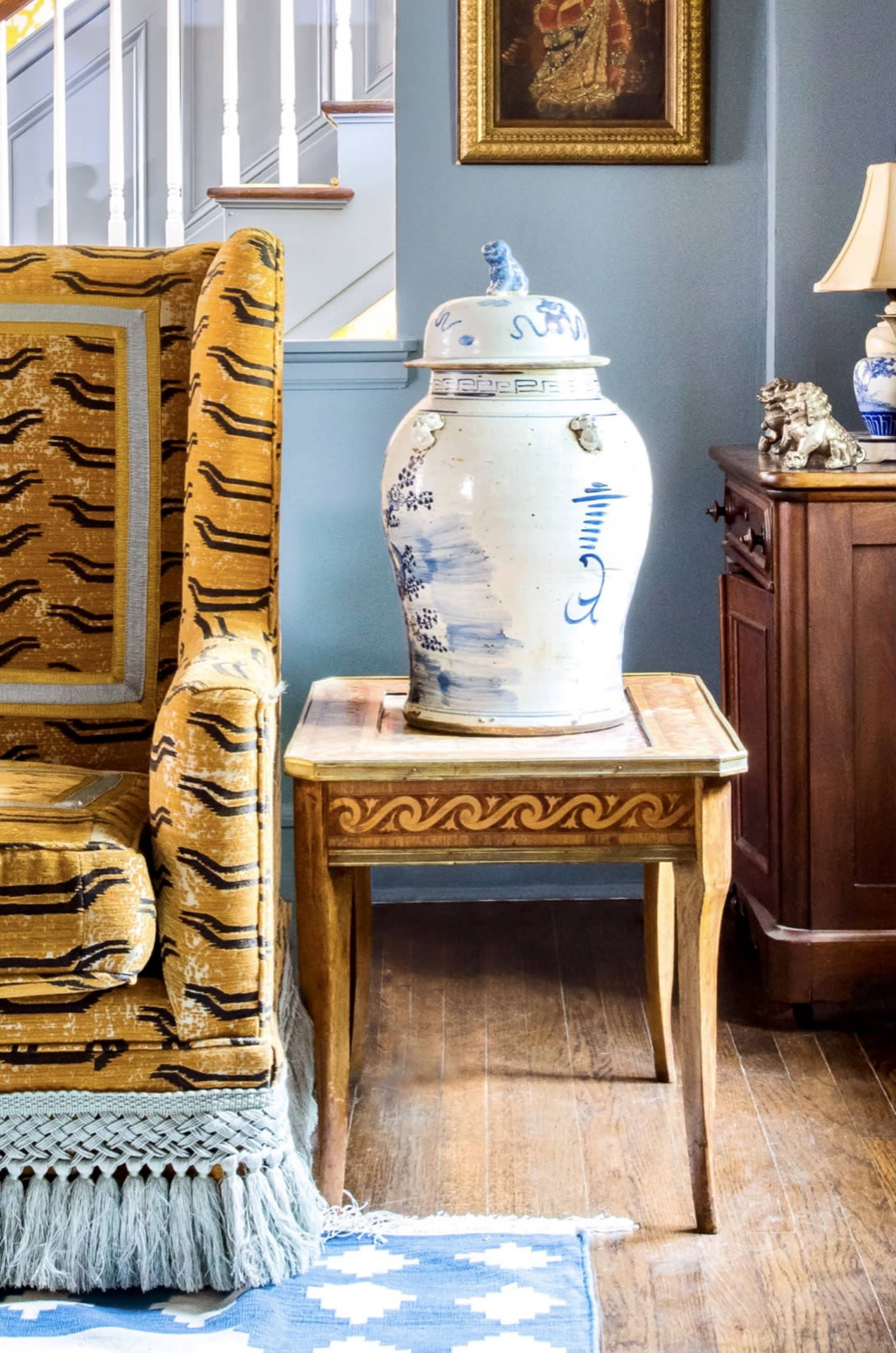A decorative ceramic jar stands on a wooden side table next to a patterned armchair in a room with wooden flooring and light blue walls.
