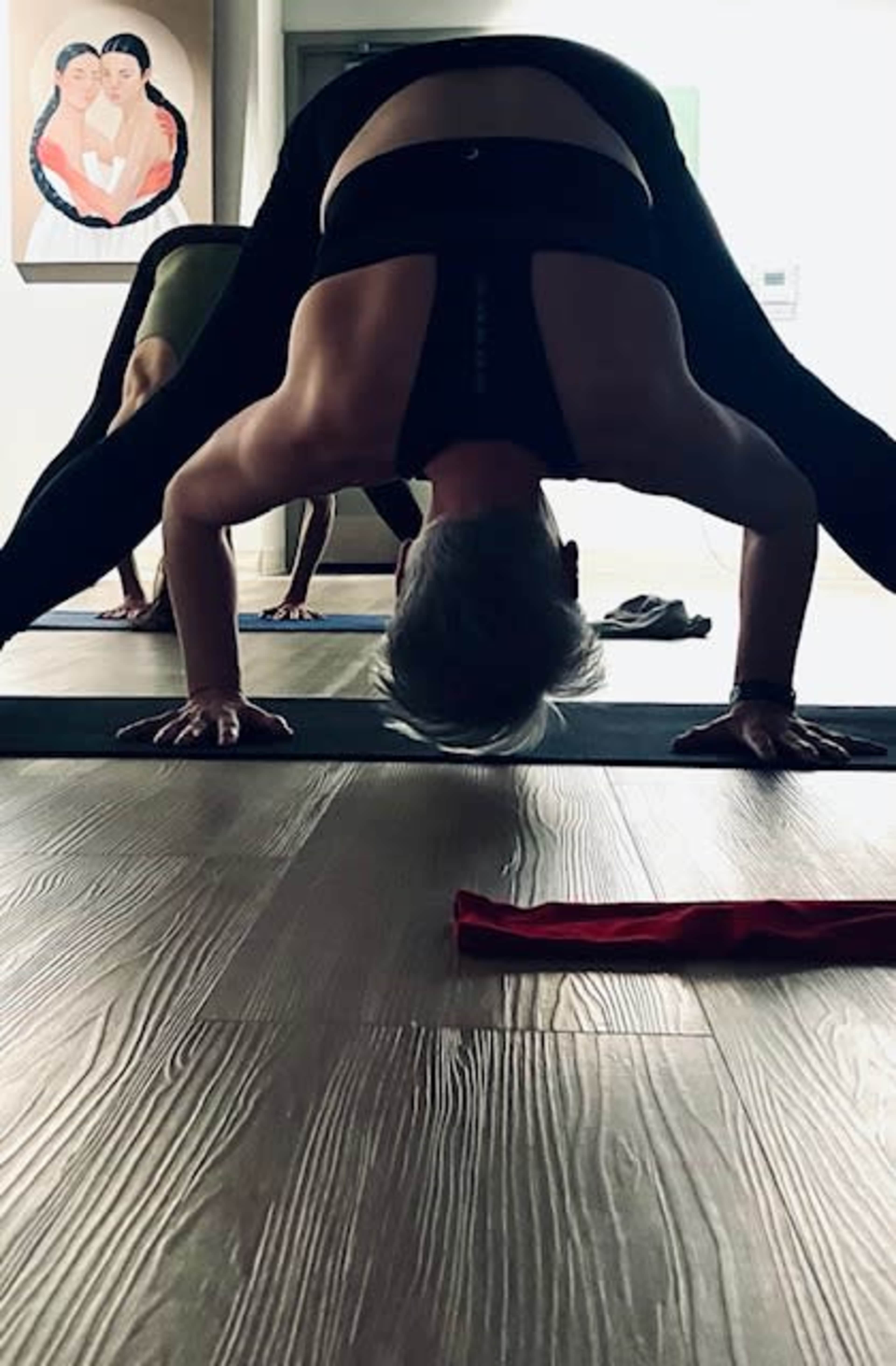 A person performs a yoga pose on a mat in a studio with another individual visible in the background.