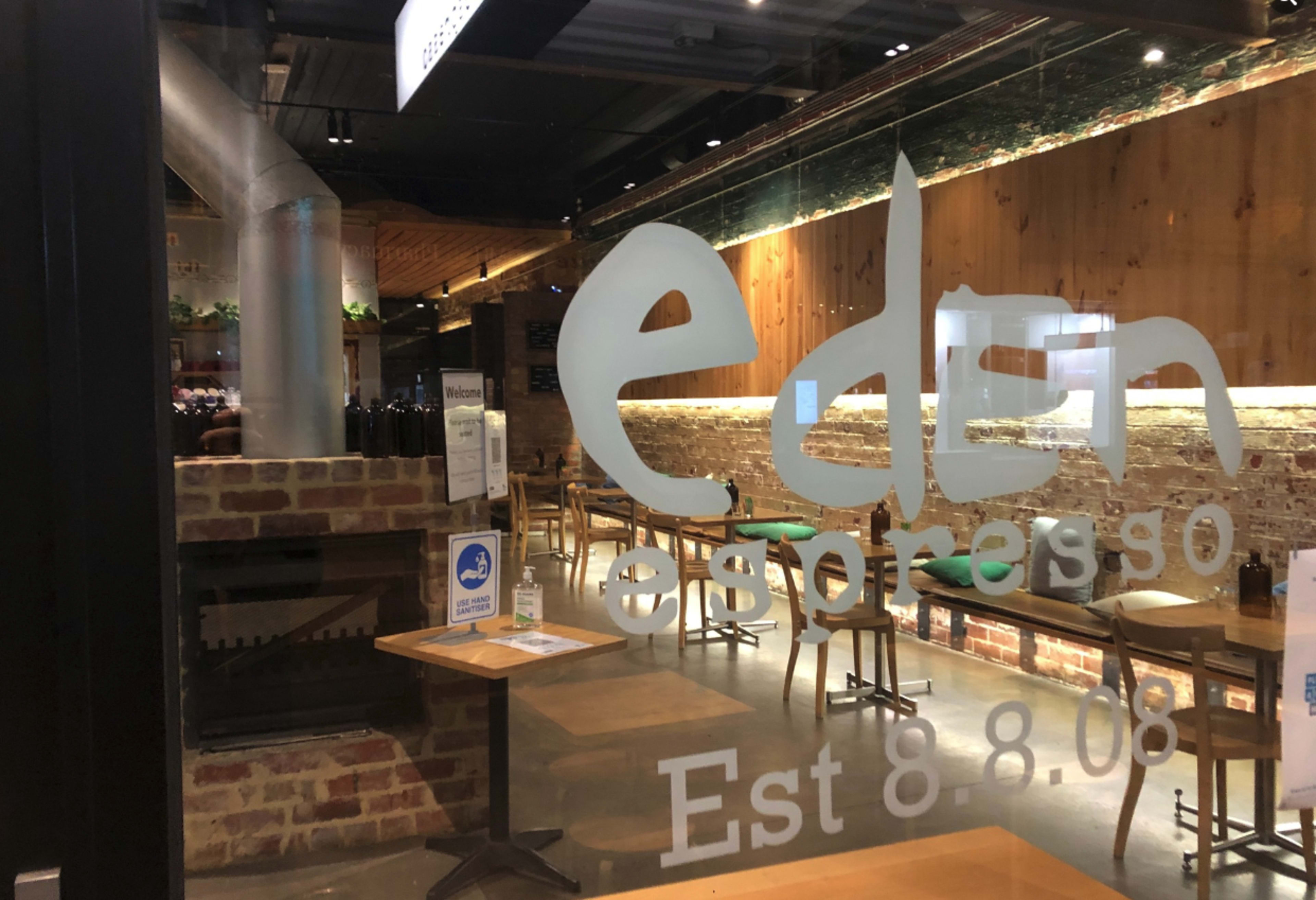 A coffee shop with wooden decor, exposed brick walls, and tables visible through a glass entrance displaying the name "eden espresso."