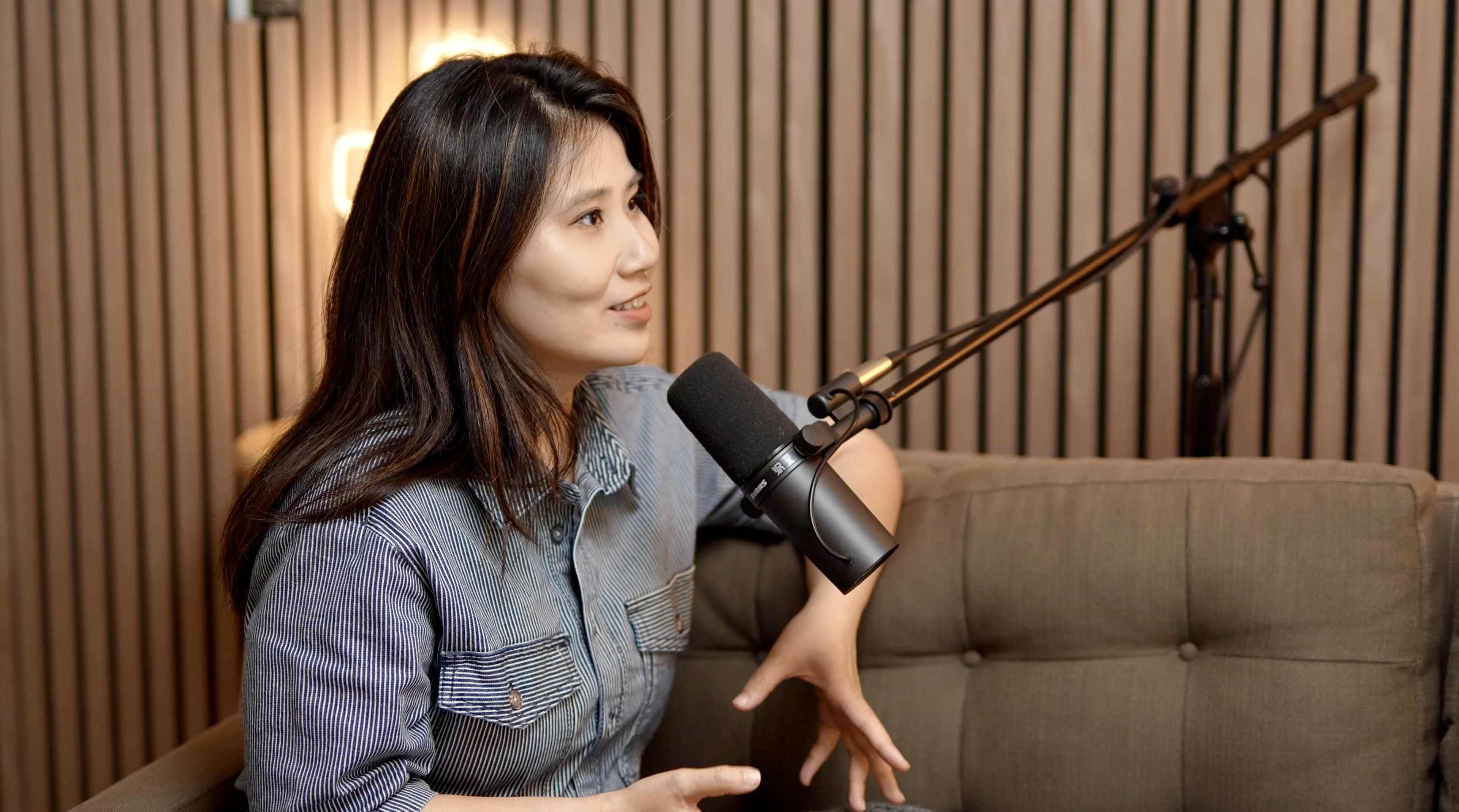 A woman with long hair is speaking into a microphone while seated on a couch in a well-lit room with wooden paneling.