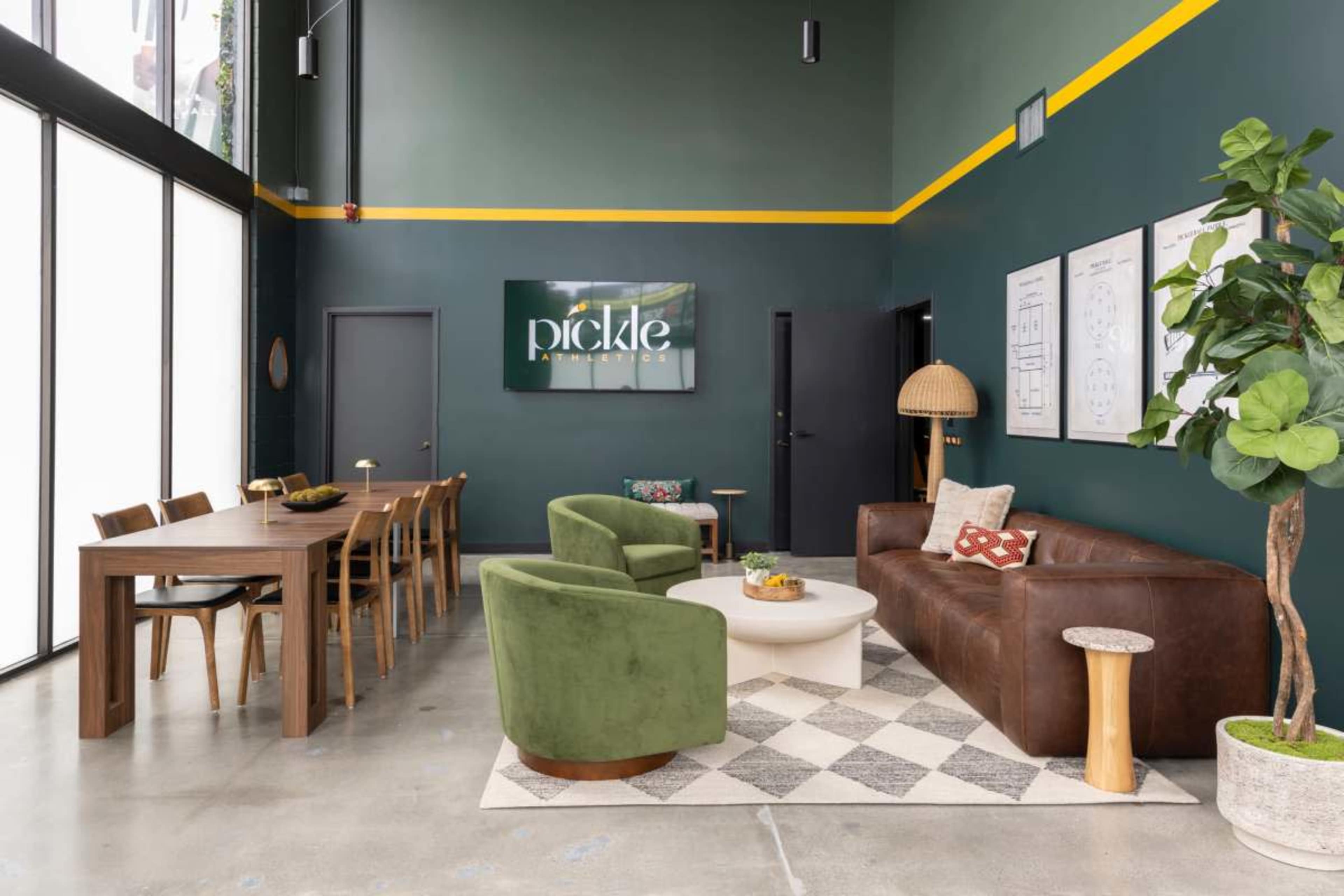 The image shows a modern interior space featuring a seating area with a brown leather sofa, green accent chairs, a white coffee table, and a long wooden dining table under a deep green wall with decorative artwork.