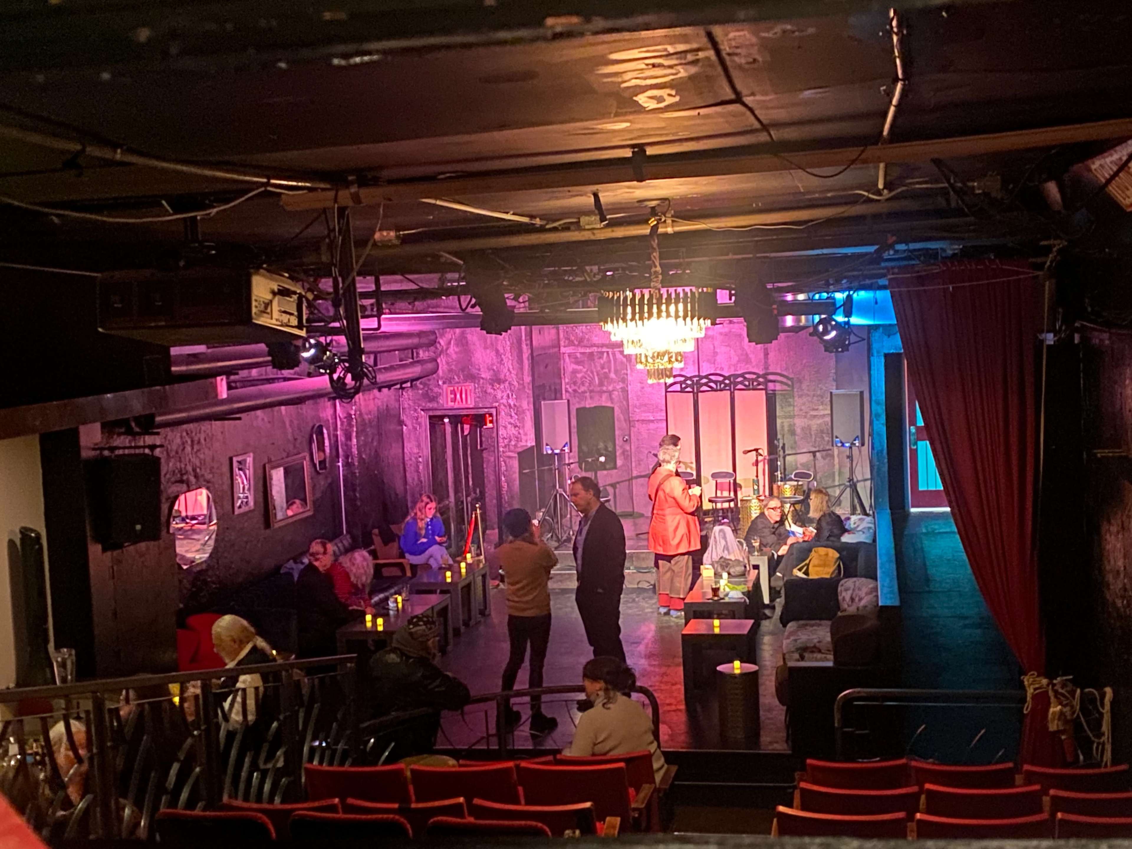 The image shows a dimly lit lounge with a small stage, seating areas, and patrons engaged in conversation.