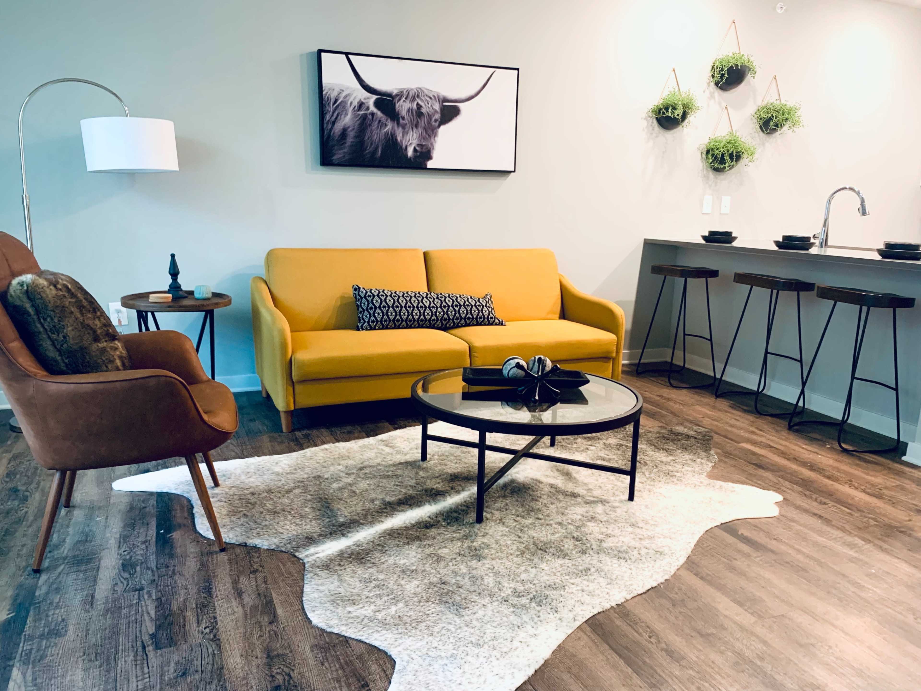 The living area features a yellow sofa, an armchair, a coffee table, and a wall-mounted artwork of a bull above a modern kitchenette with bar stools.