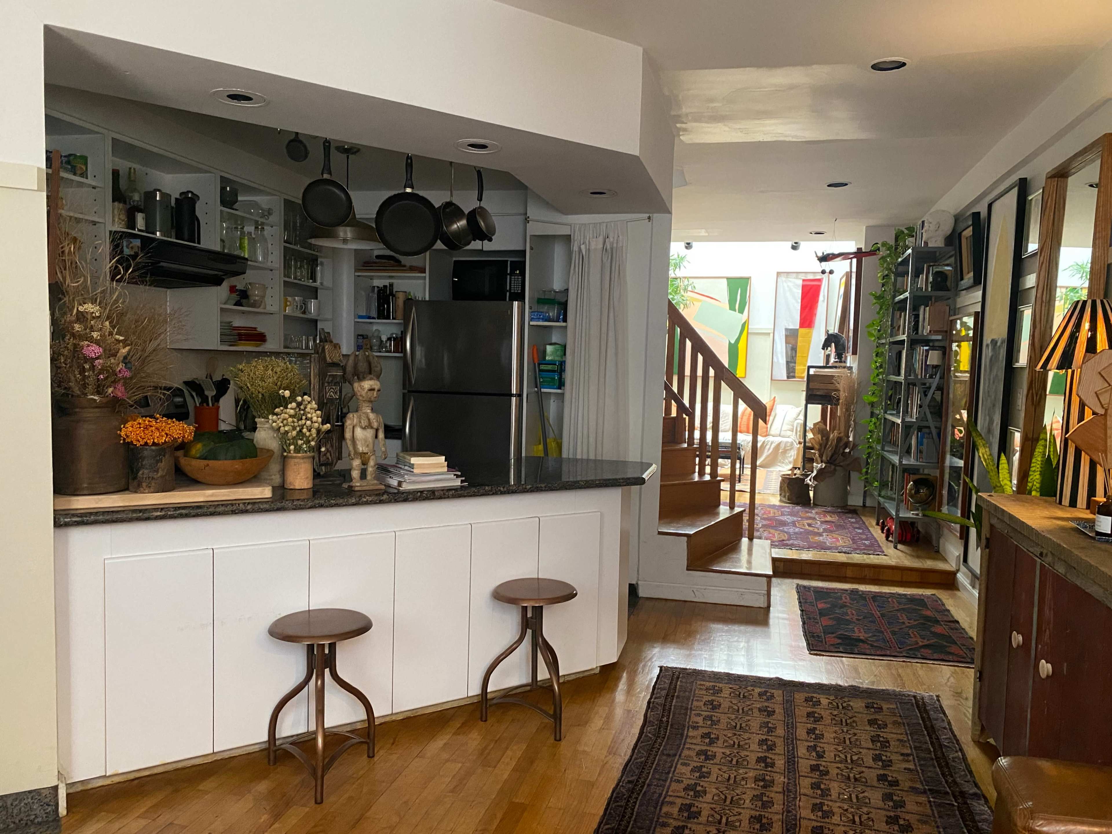 The image shows a modern kitchen with a central island, two stools, and an open living space adorned with various decorations and plants.