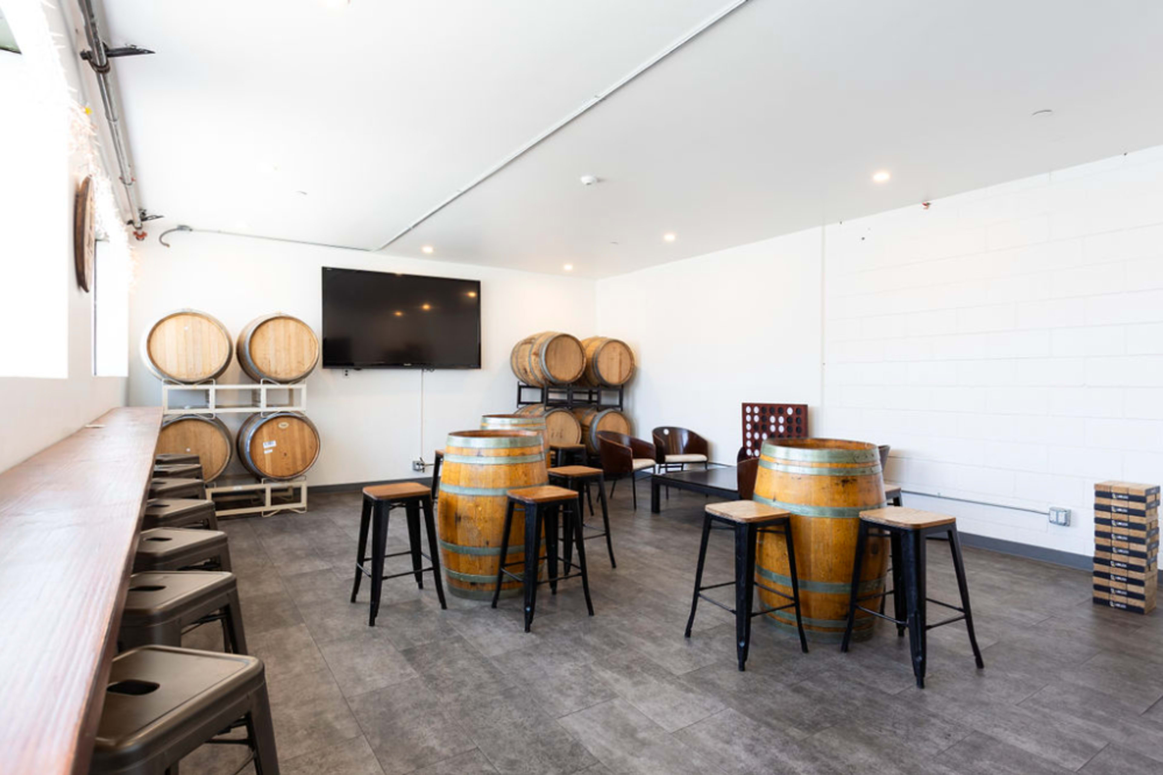 The room features wooden barrels stacked against a wall, a large flat-screen TV, and a seating area with tables and chairs.