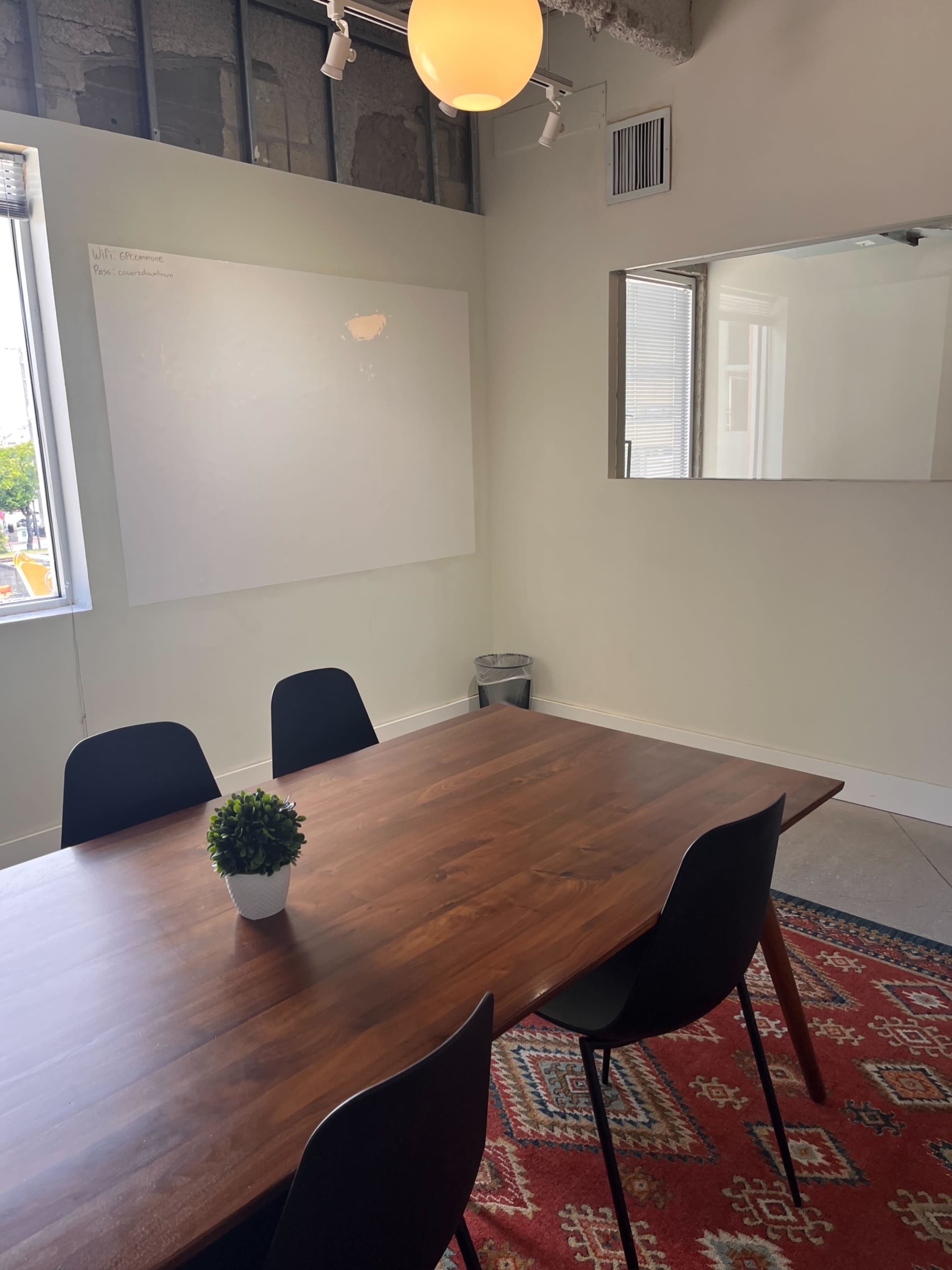 The image shows a small meeting room featuring a wooden table surrounded by black chairs, a potted plant on the table, a whiteboard on the wall, and a mirror reflecting part of the room.