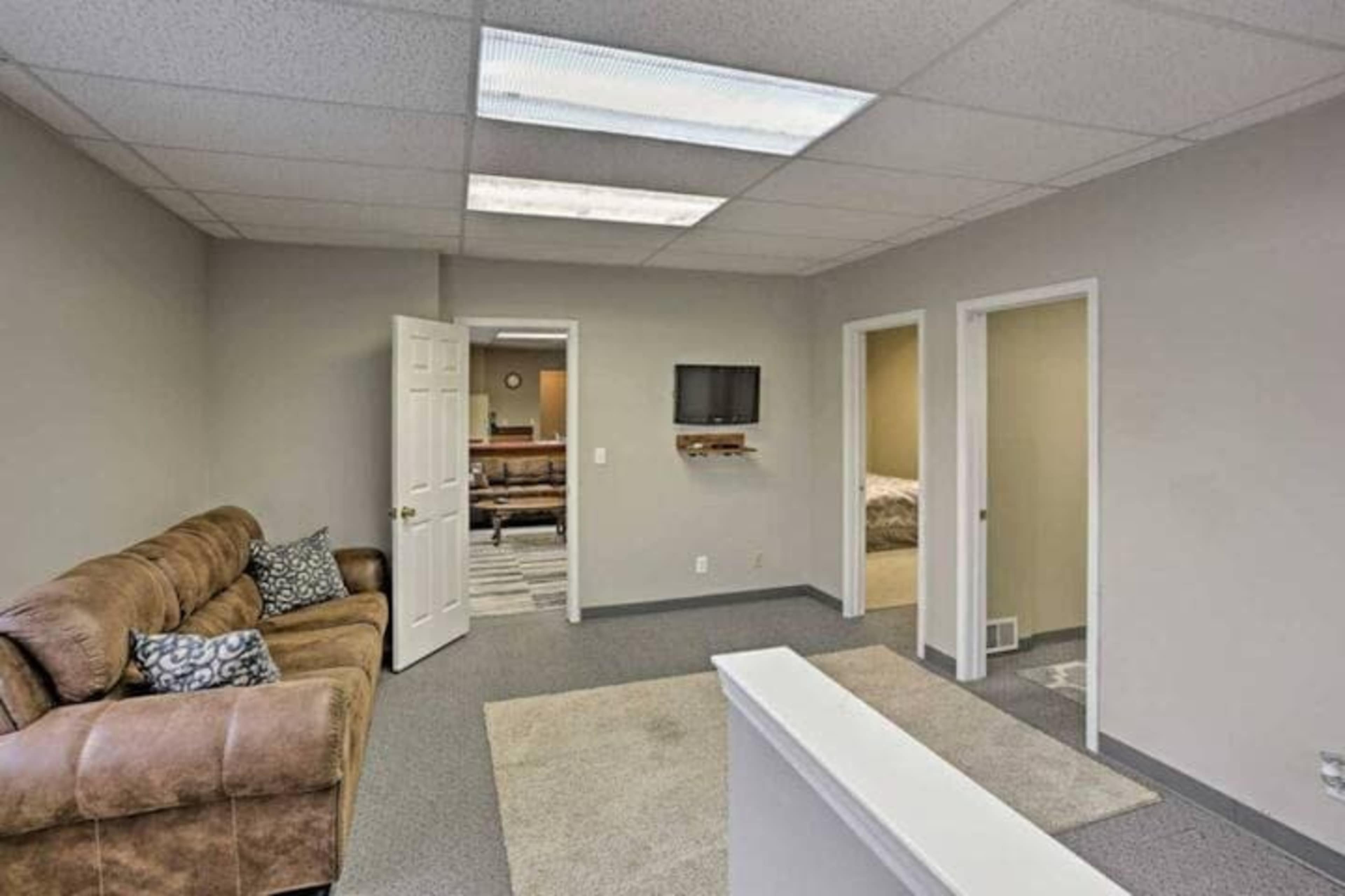 A small office space featuring a brown couch, a light-colored carpet, and two closed doors leading to other rooms, with a television mounted on the wall.