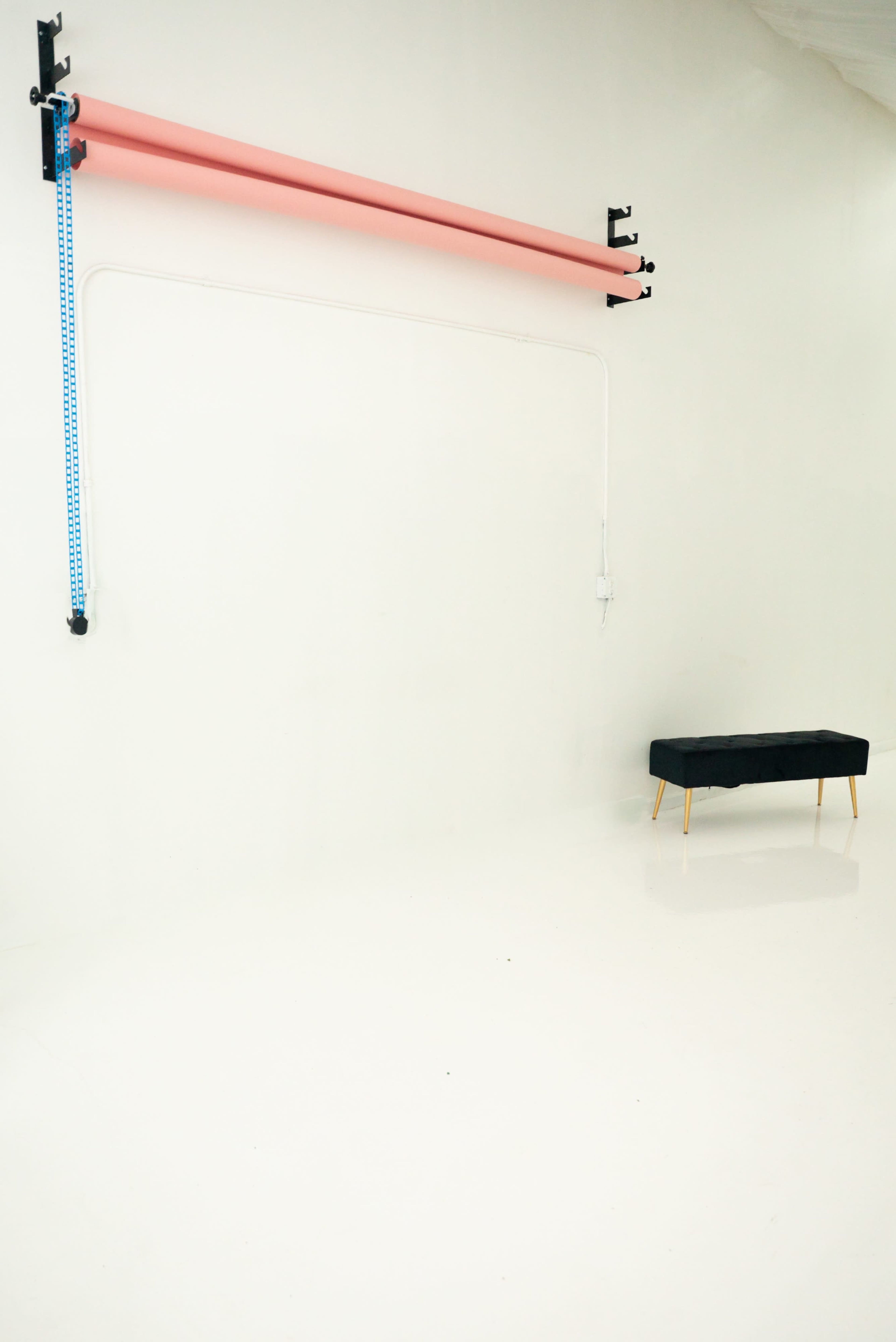 A blank photography studio features a pink backdrop hanging from a rail and a black bench against a white wall.