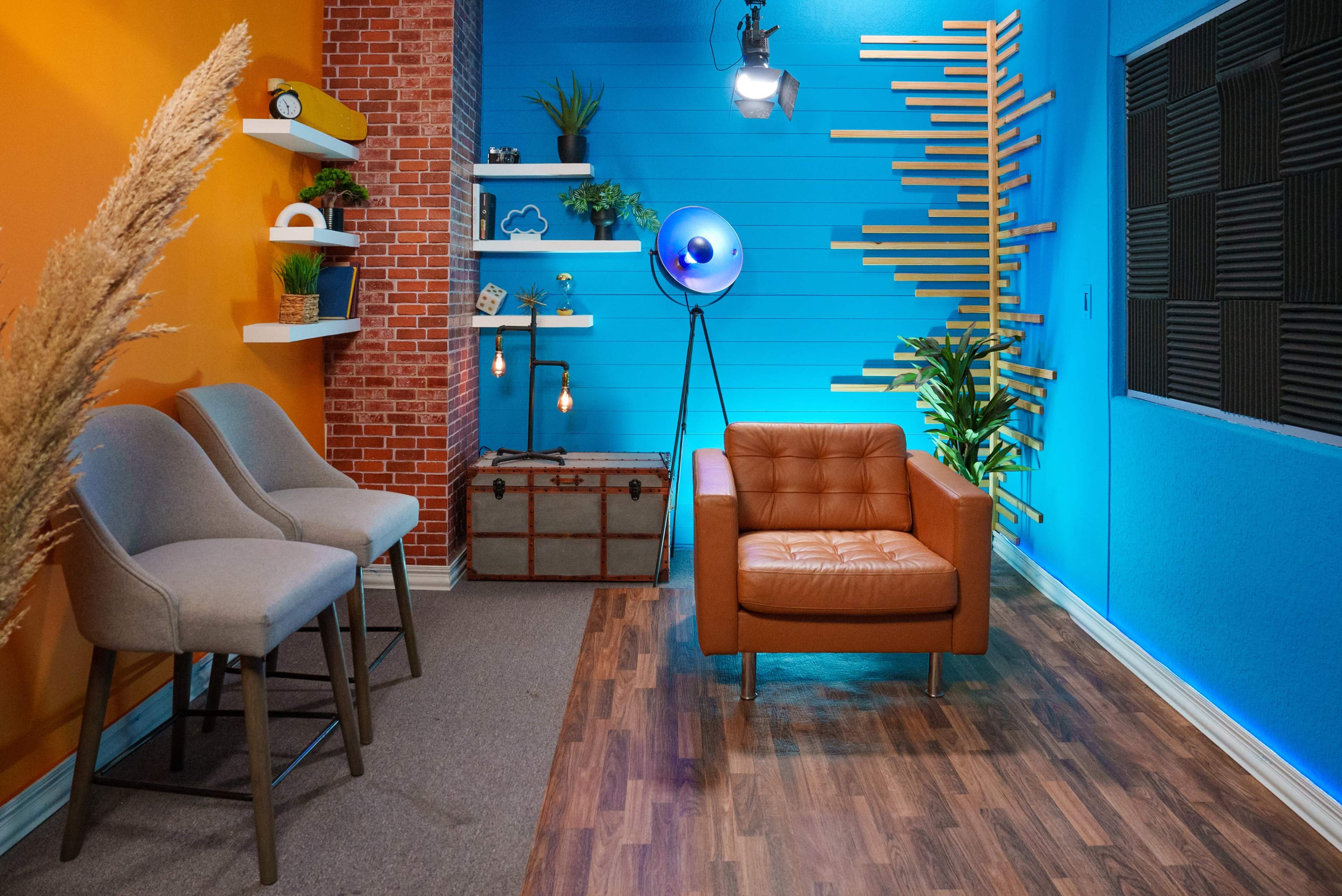 The image shows a styled interior space featuring a brown leather chair, three light-colored bar stools, various wall shelves with decorative items, and a blue and orange color scheme.