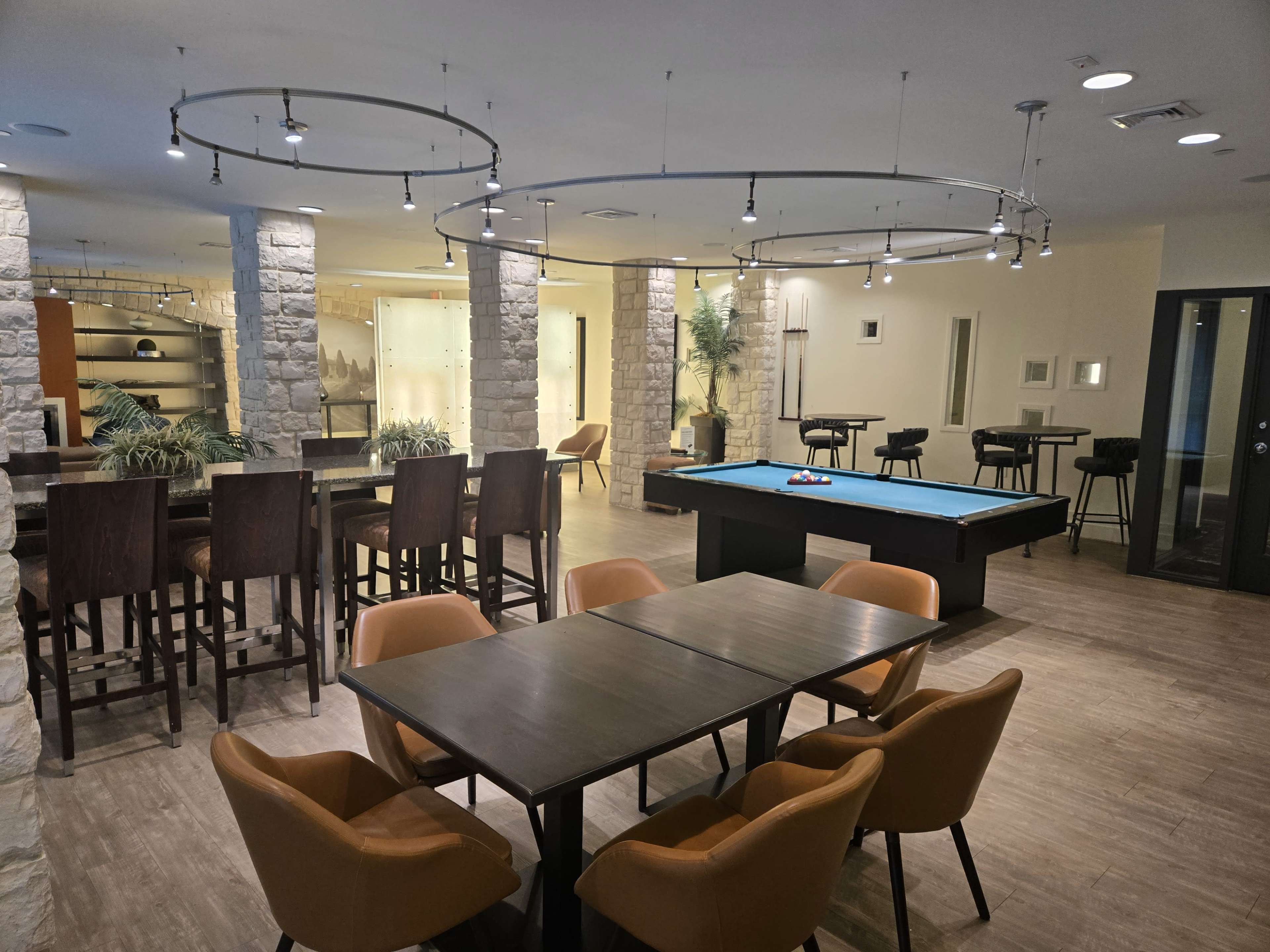 The image shows a modern leisure area featuring a pool table, high tables with stools, and a dining table surrounded by orange chairs.