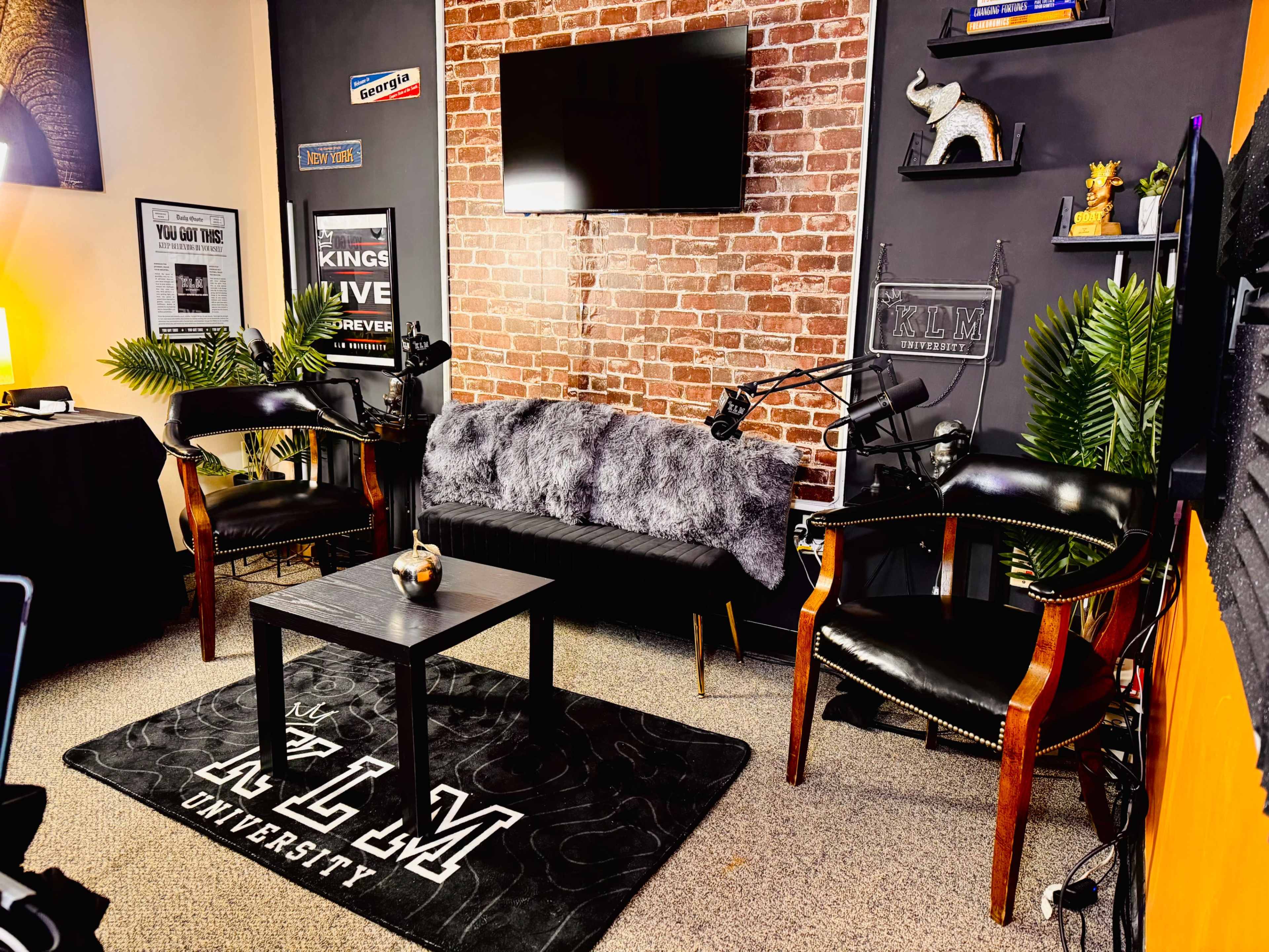 The image depicts a modern interview or podcast studio featuring a brick wall backdrop, a sofa with throw blankets, two chairs, a coffee table, and various decorative elements.