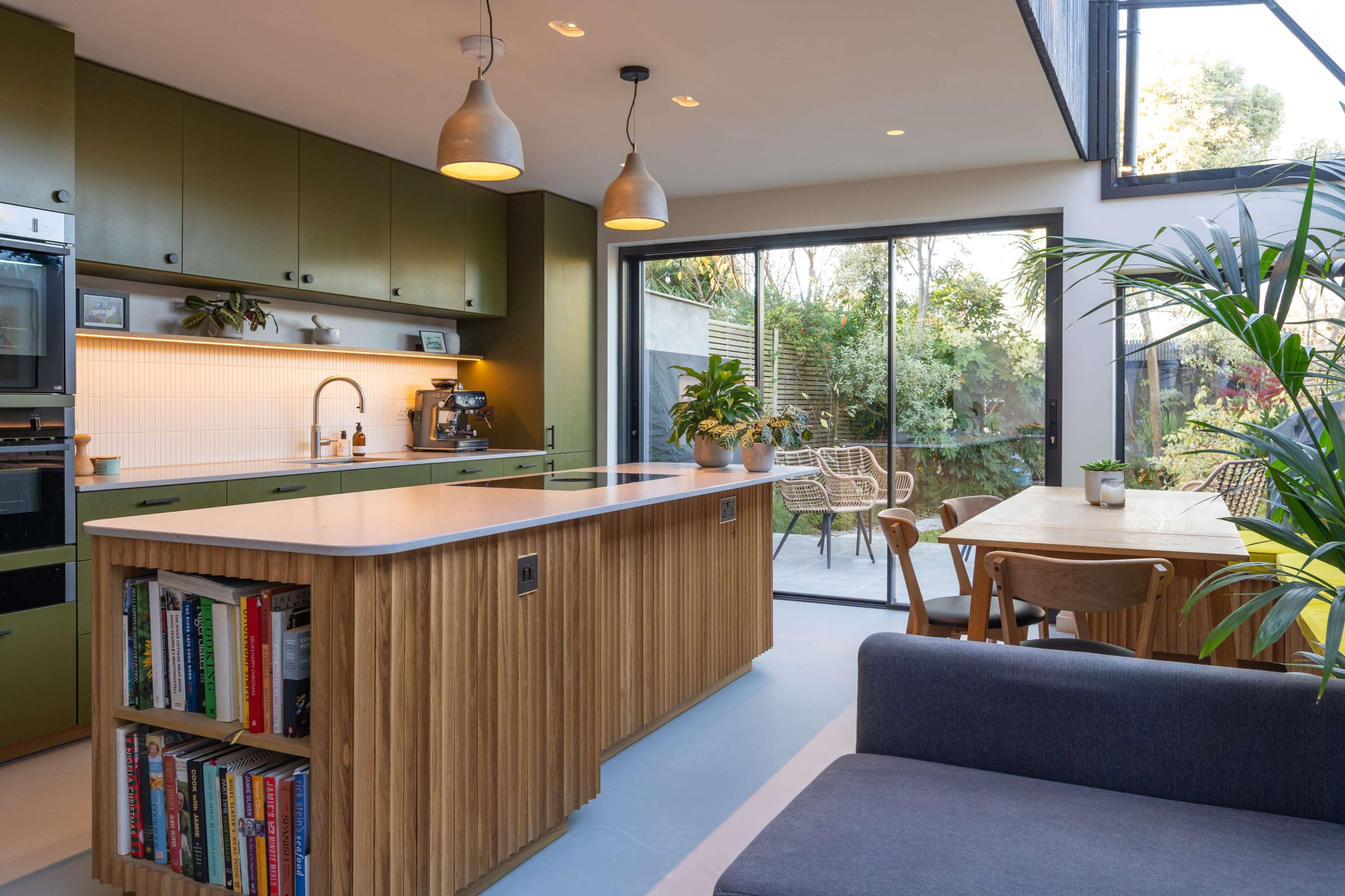 The image shows a modern kitchen with green cabinets, a wooden island featuring a bookshelf, and large windows overlooking a garden.