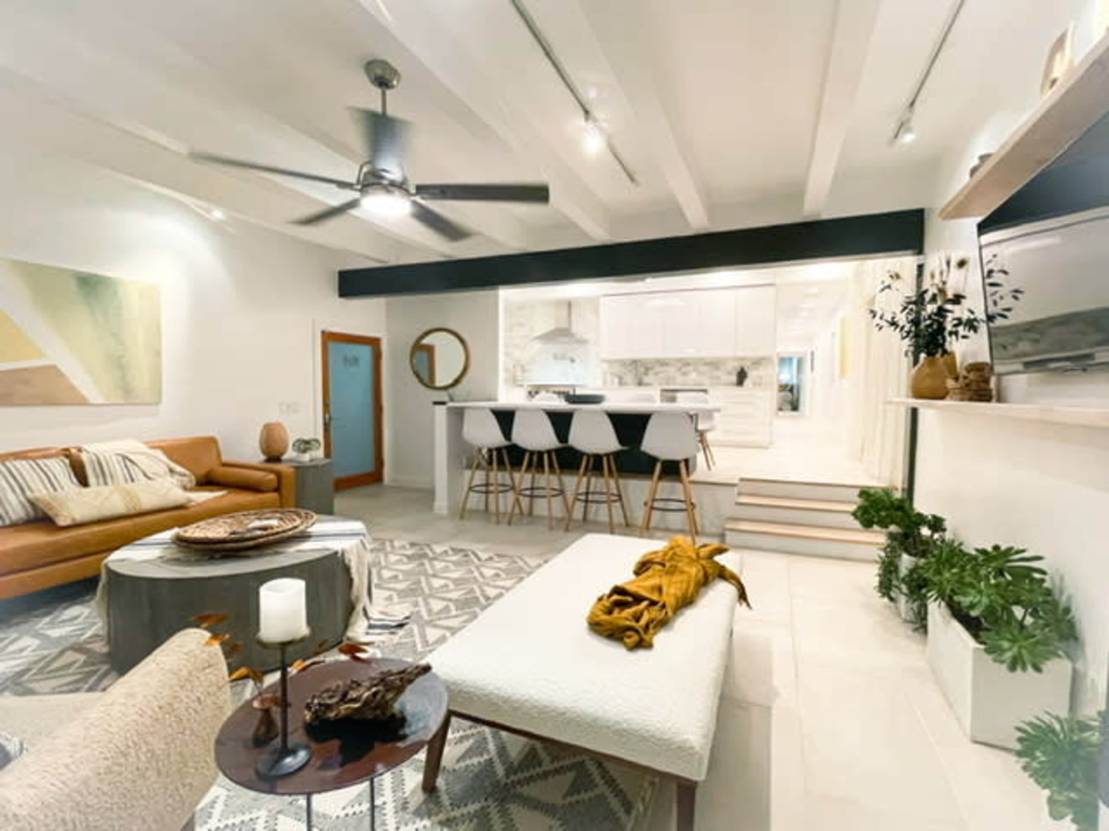 A modern living room features a brown sofa, a large area rug, a round coffee table, and a view of a kitchen with white cabinetry and bar seating.