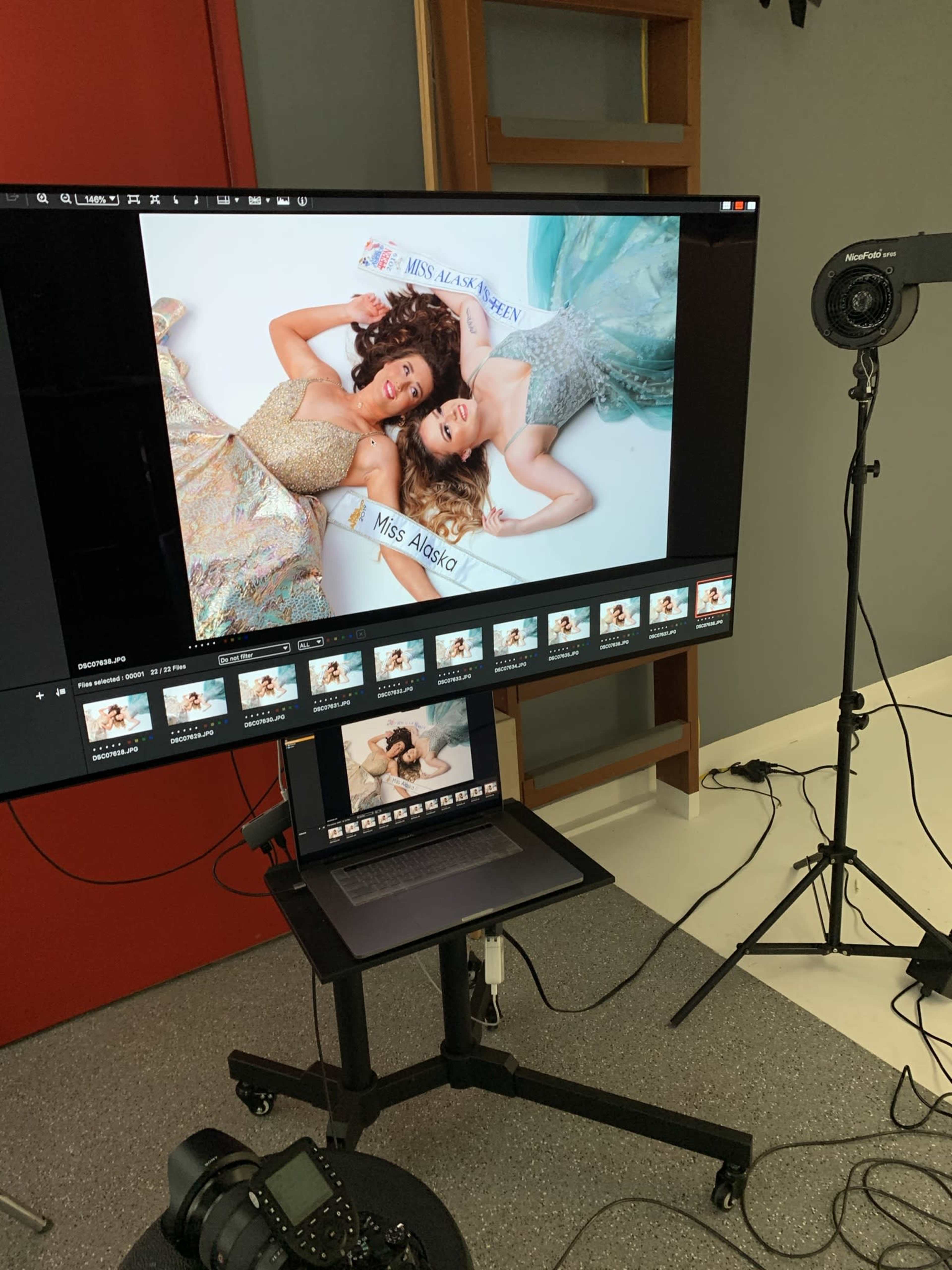 Two women in formal dresses posing for a photoshoot, with a computer and monitor displaying the images taken.