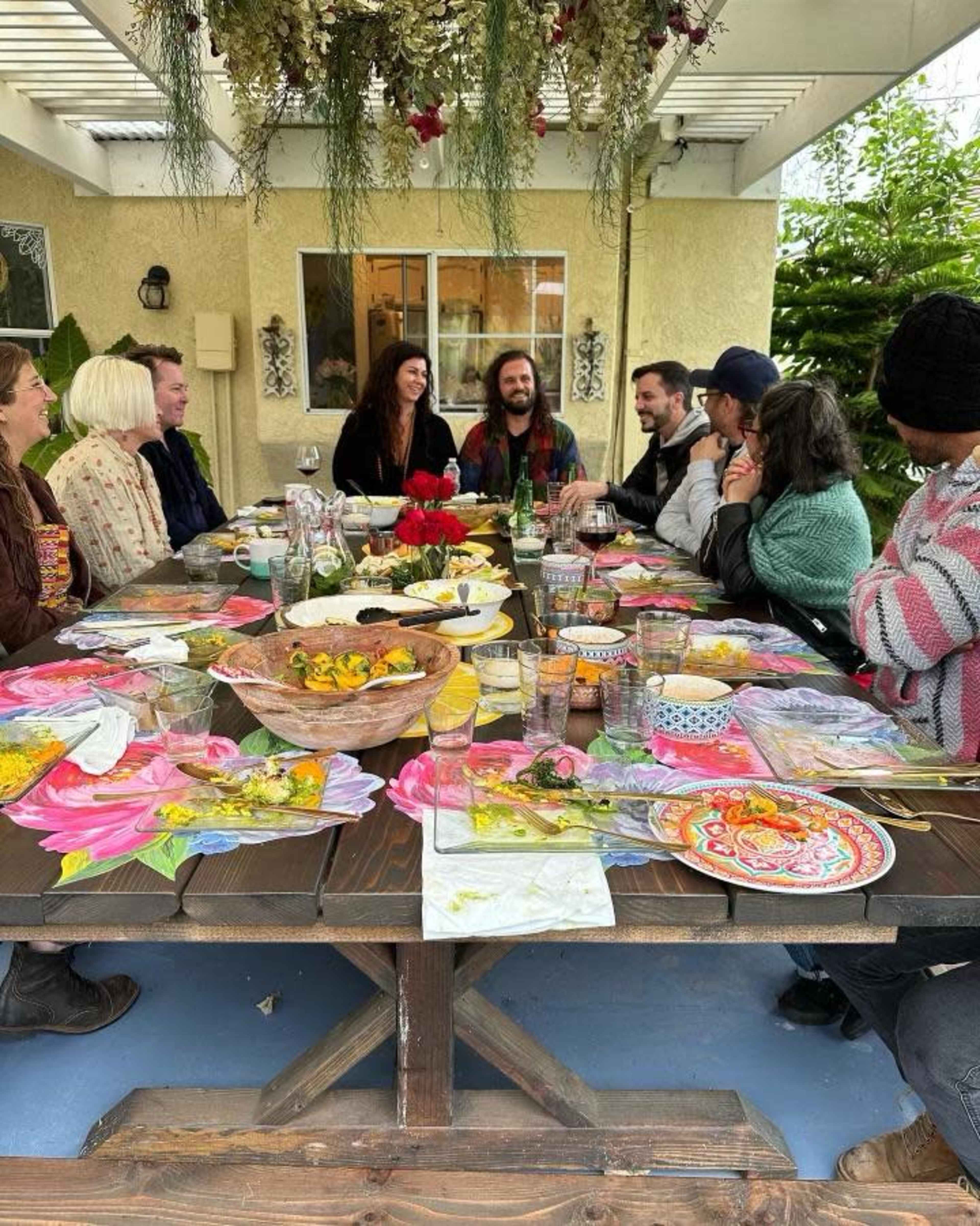A group of people sits around a large wooden table adorned with colorful plates and various dishes for a meal outdoors.