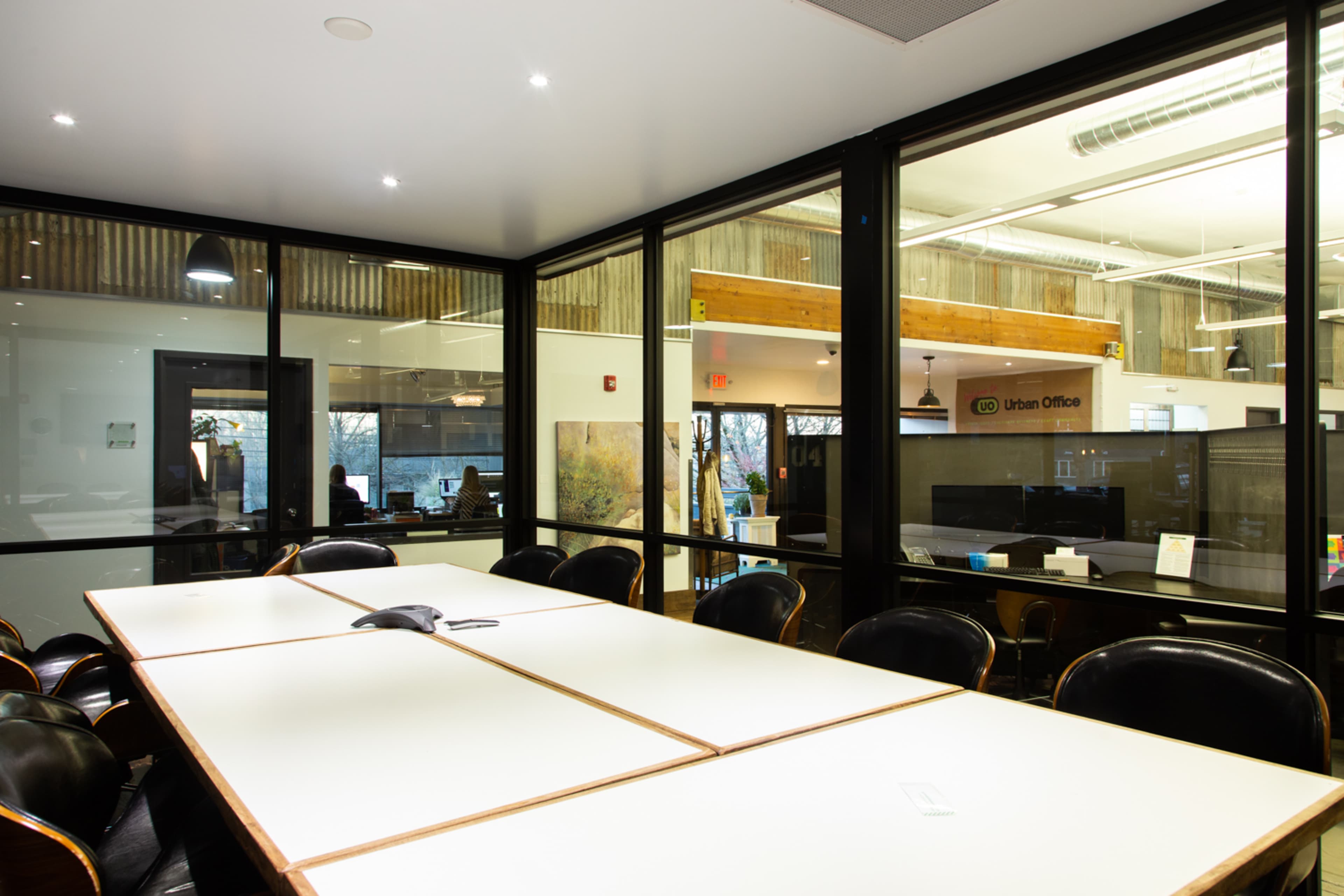 A modern conference room with a large table and glass walls, overlooking a workspace with computer stations and wooden accents.