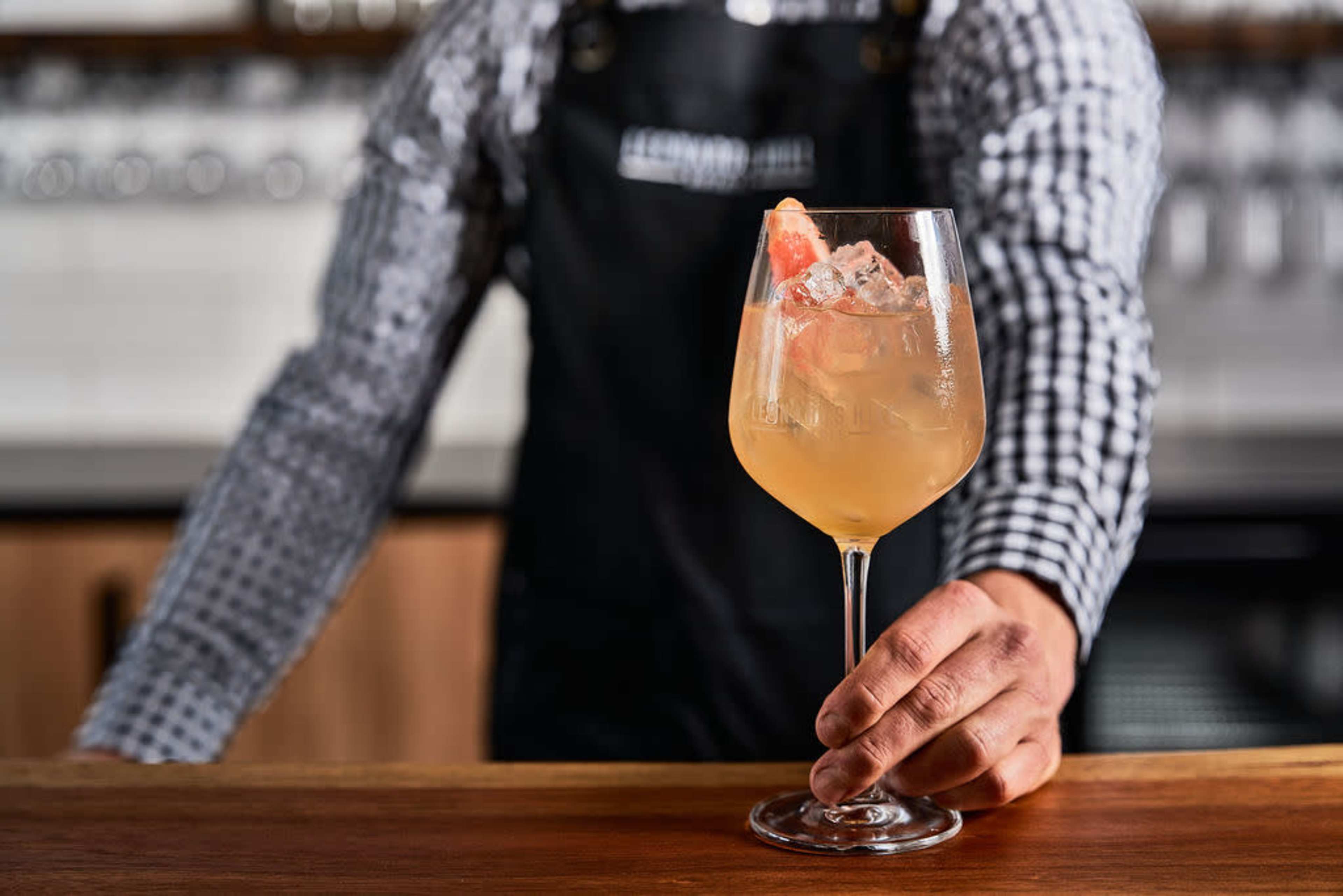 A bartender in a checked shirt and black apron holds a cocktail glass filled with a light-colored drink garnished with ice and a slice of grapefruit.