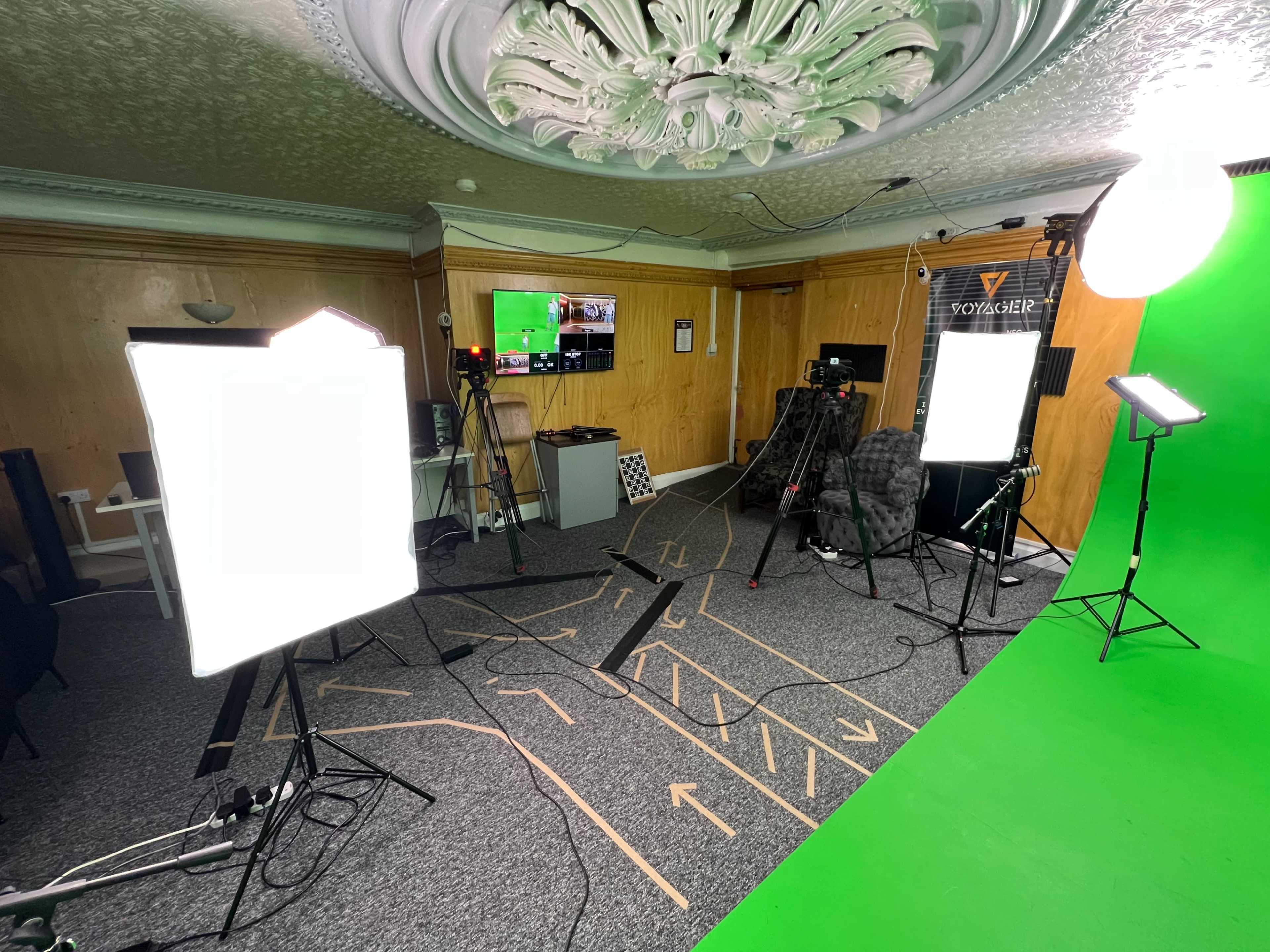 The image shows a studio setup with multiple cameras, lighting equipment, and a green screen, all arranged in a room featuring wood paneling and a decorative ceiling.