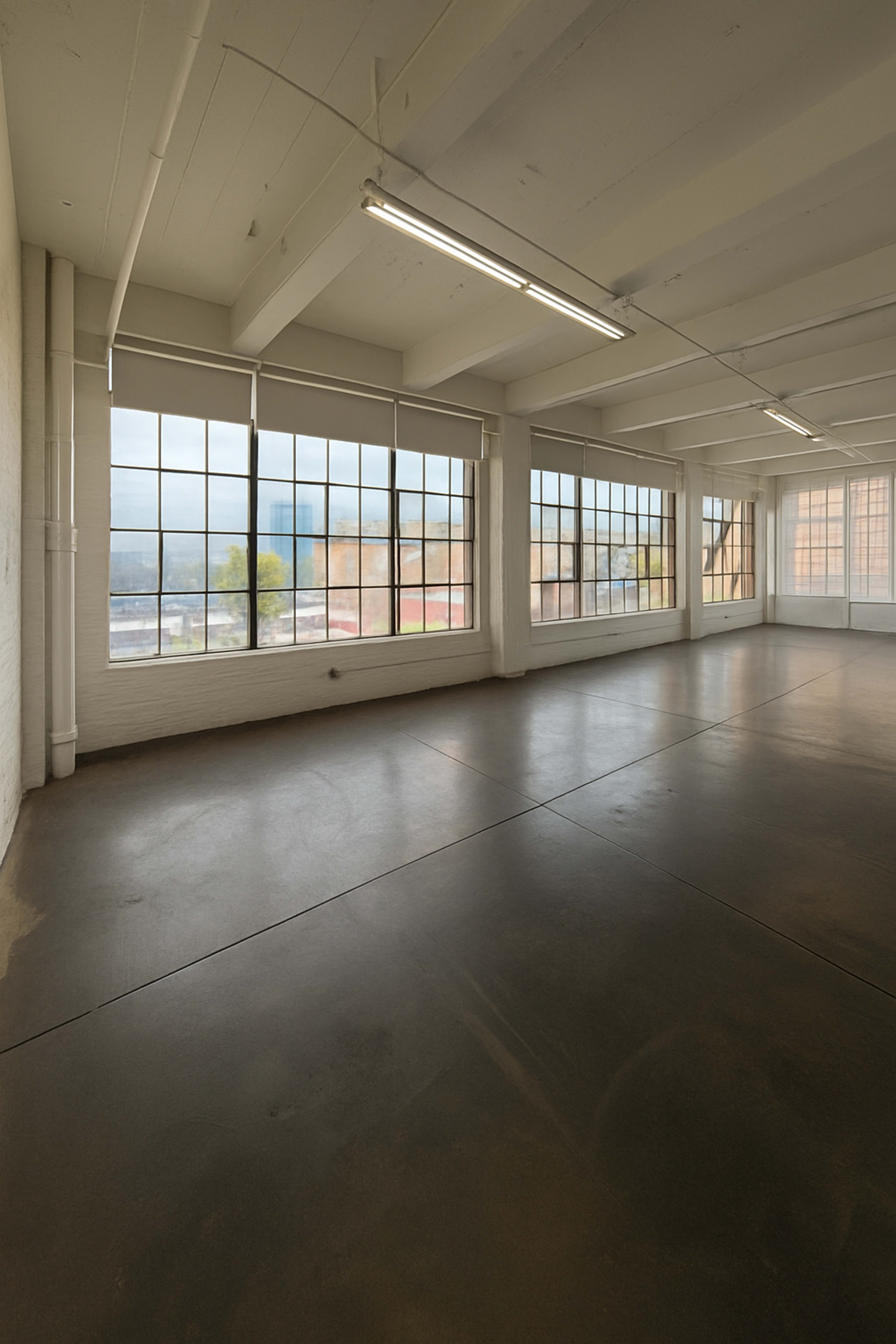 The image shows a spacious, empty room with large windows along one wall and concrete flooring.
