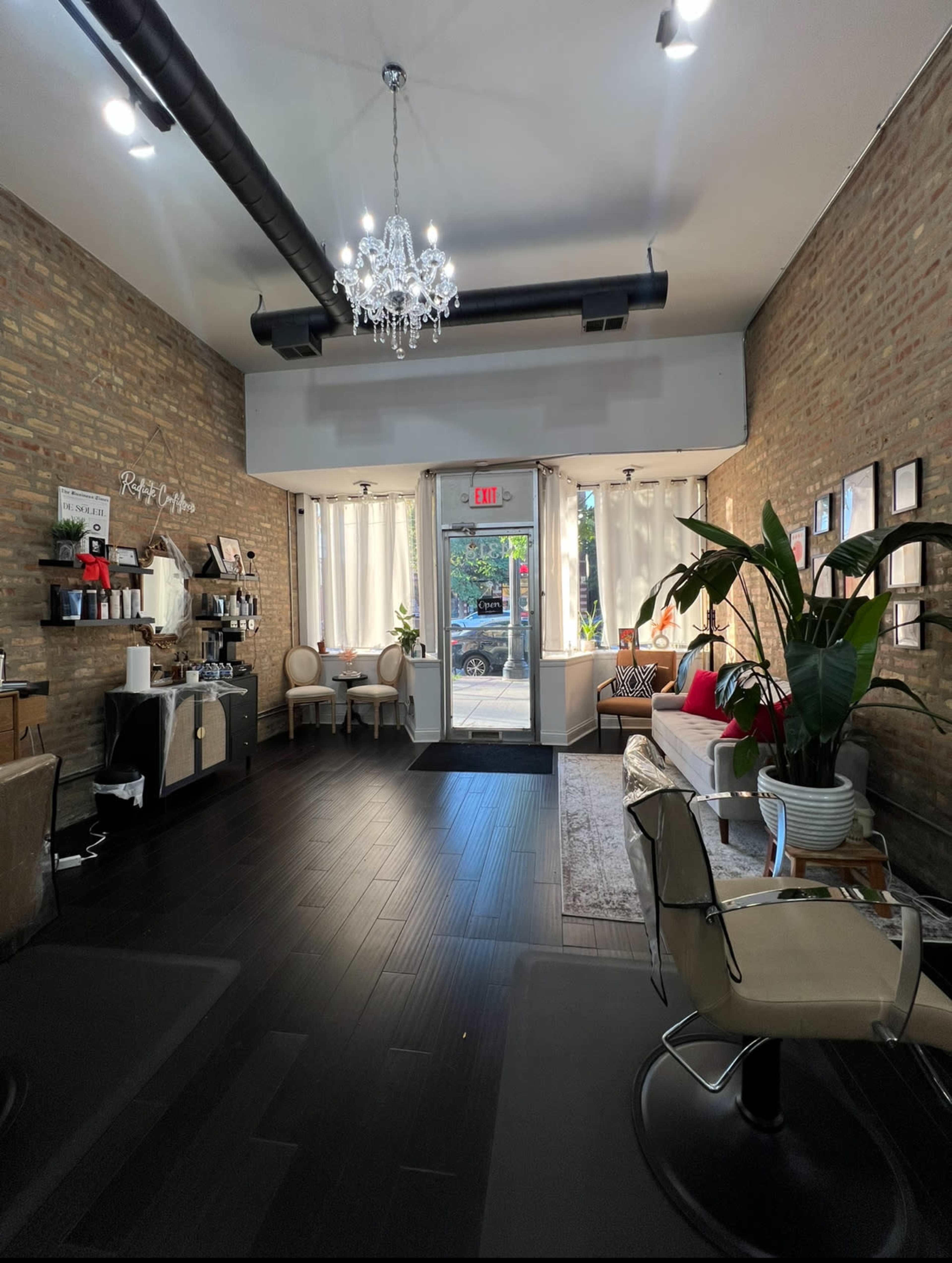 The image shows a stylish interior of a salon or spa featuring a seating area, a coffee station, large windows, and decorative plants.
