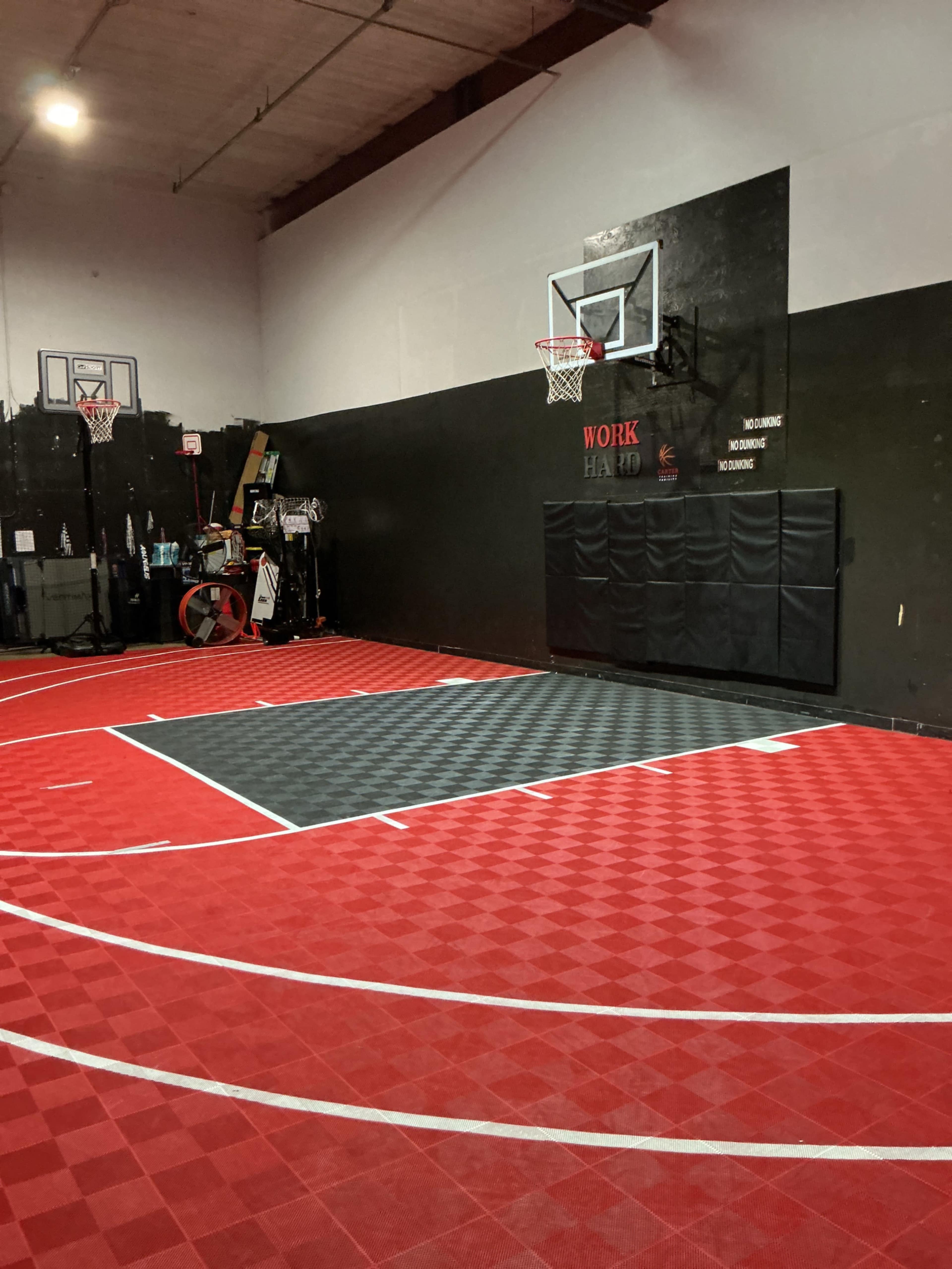 The image shows an indoor basketball court featuring red and gray flooring, two basketball hoops, and a black wall with the word "WORK" displayed.