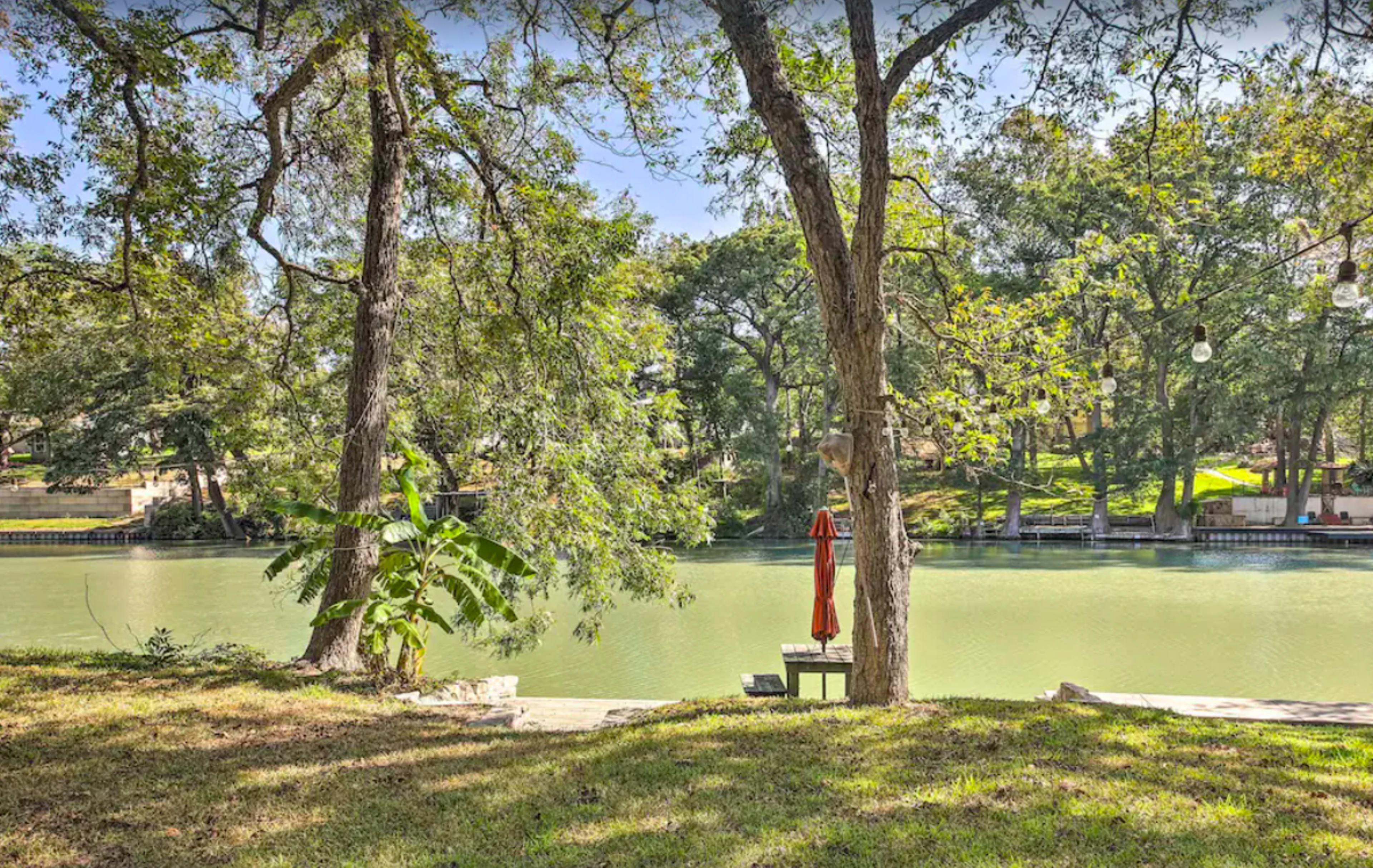 The scene shows a calm river bank framed by trees, with a lone chair and an umbrella positioned near the water's edge.