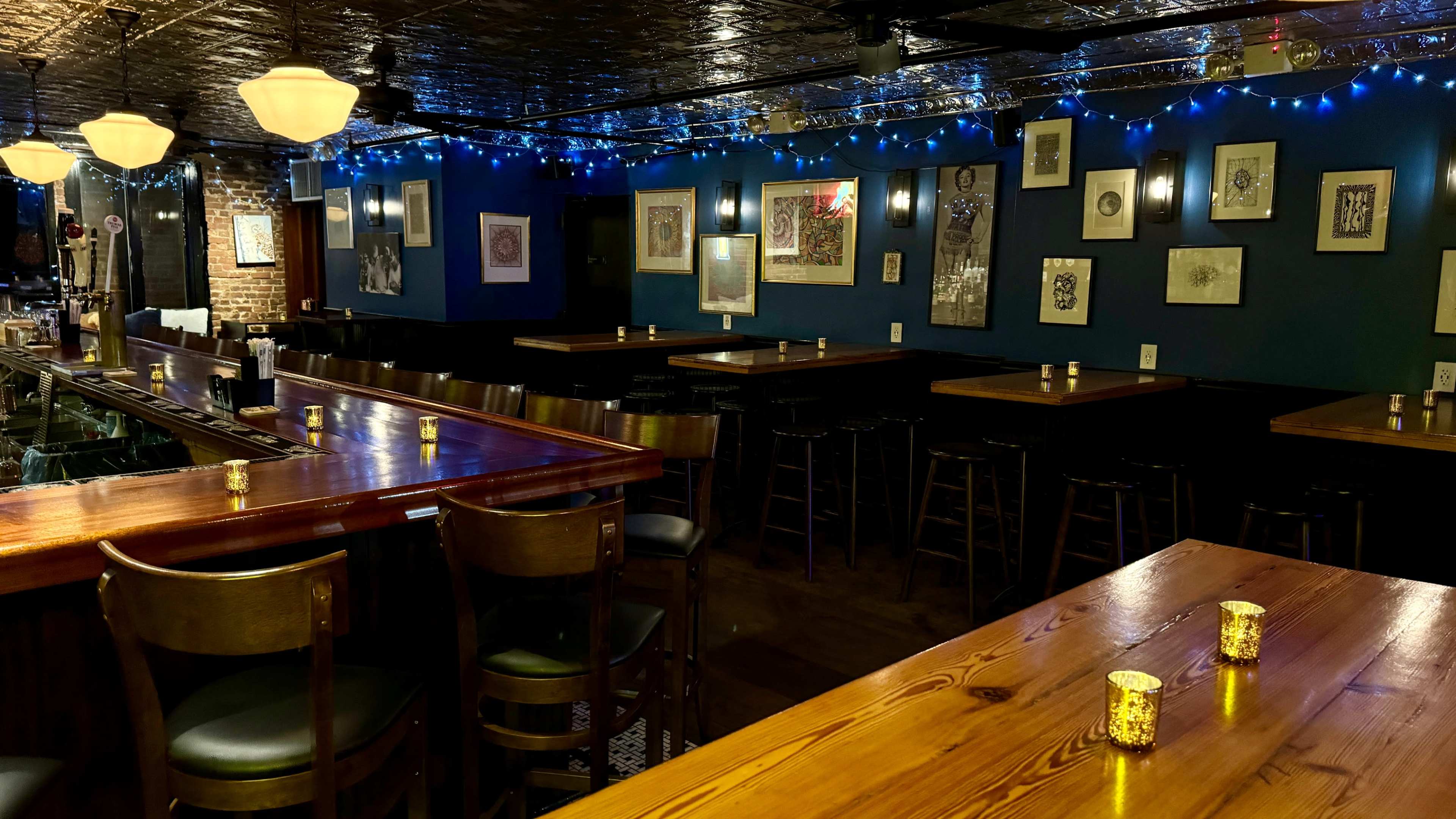 An empty bar area with wooden tables and chairs, adorned with framed artwork and blue string lights.