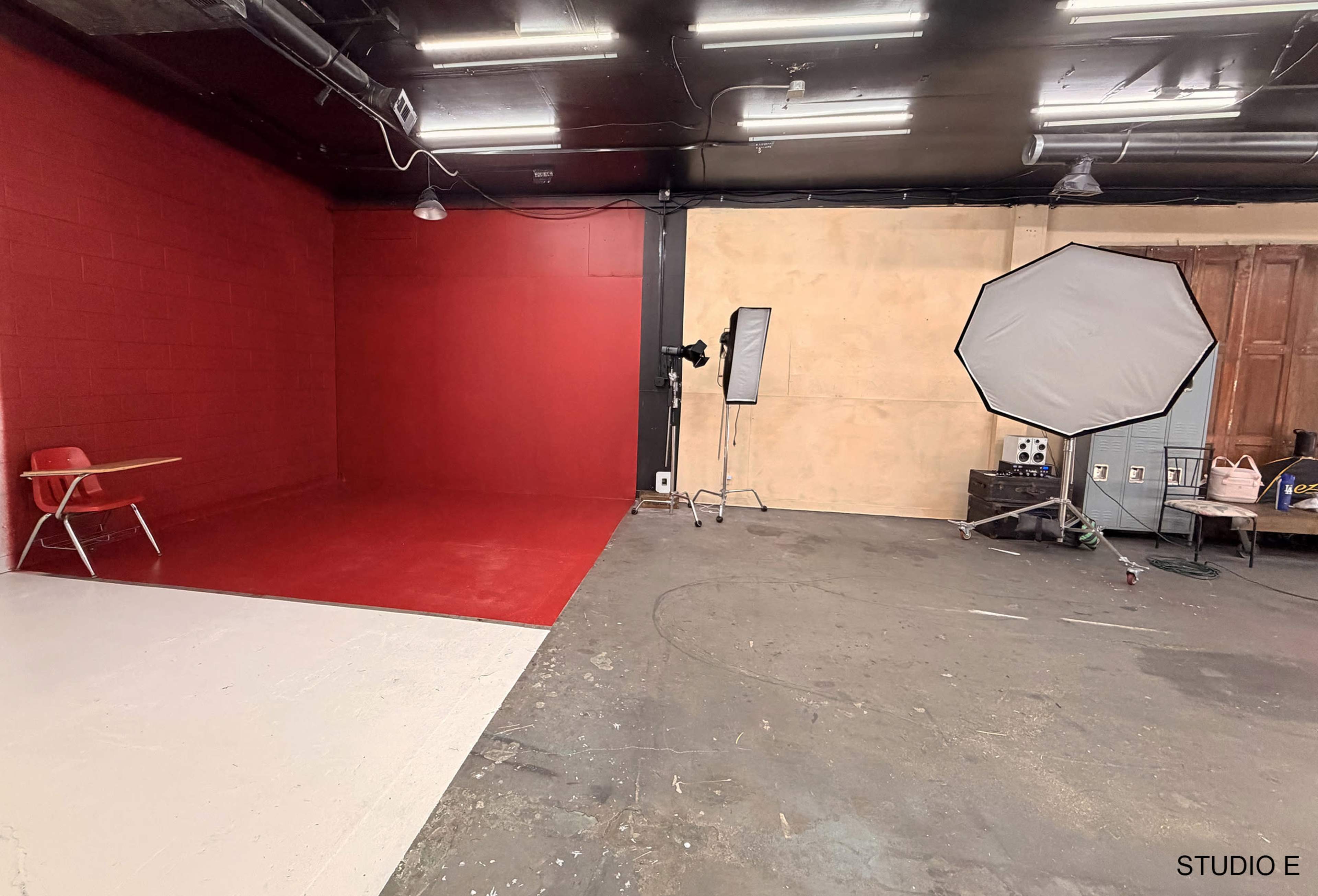 The image shows a photography studio featuring a red wall and a white floor on one side, with a chair and lighting equipment set up in the space.