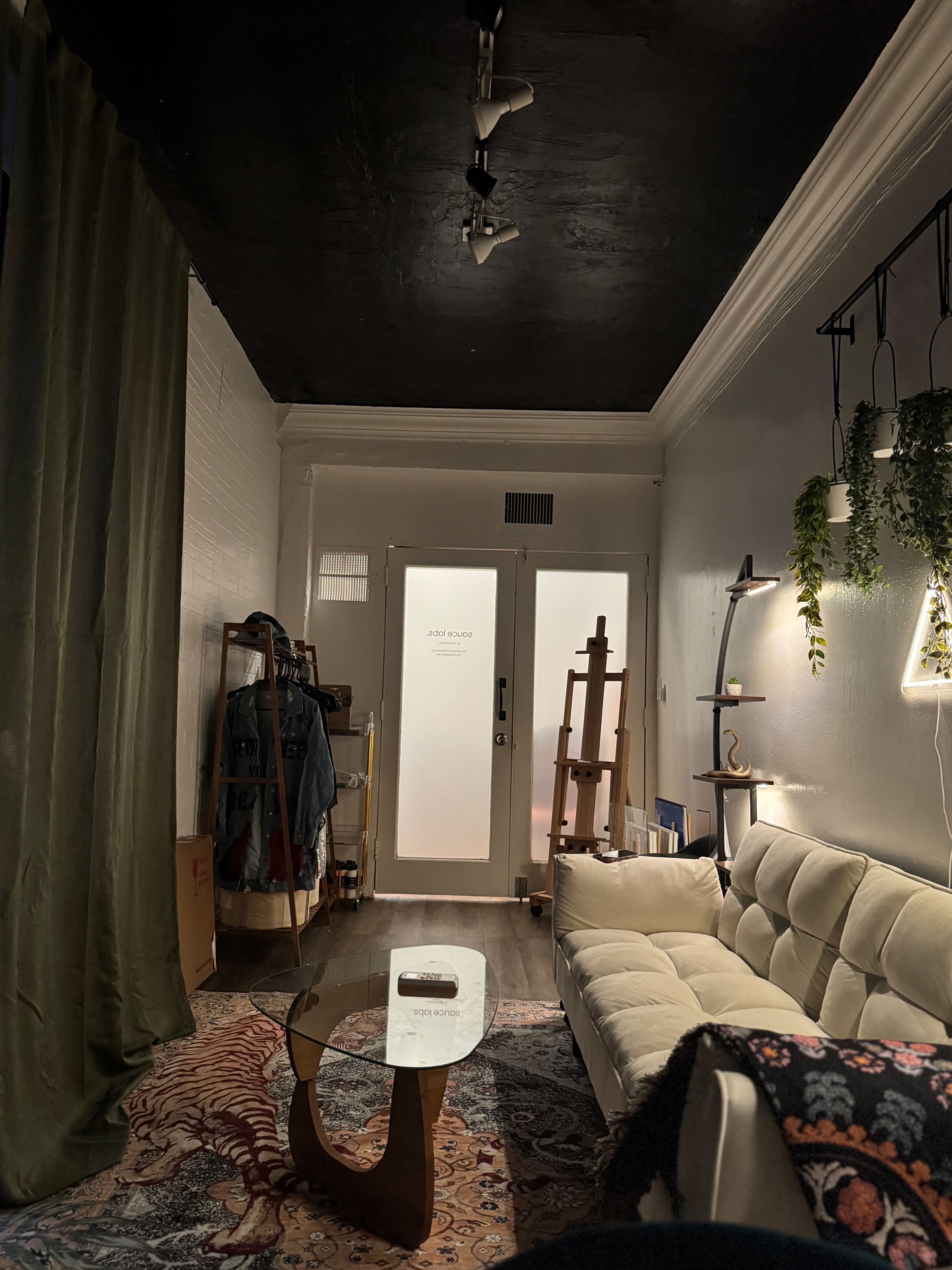 The image shows a cozy living space featuring a white couch, a glass coffee table, an easel, and various decorative elements, with a door leading outside.