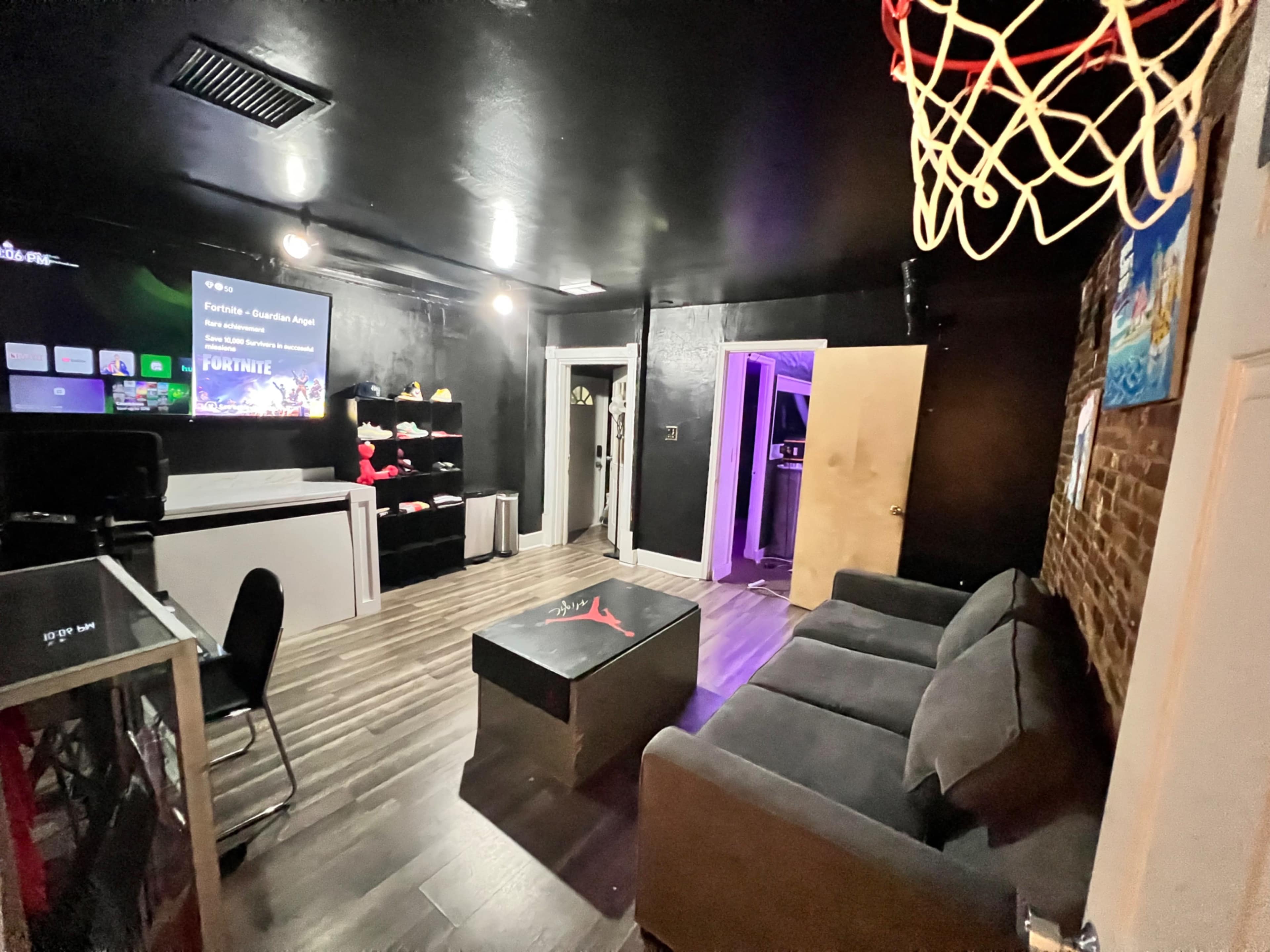 A recreation room with a gaming setup, a basketball hoop, a gray couch, and a visible wall featuring a Fortnite screen.