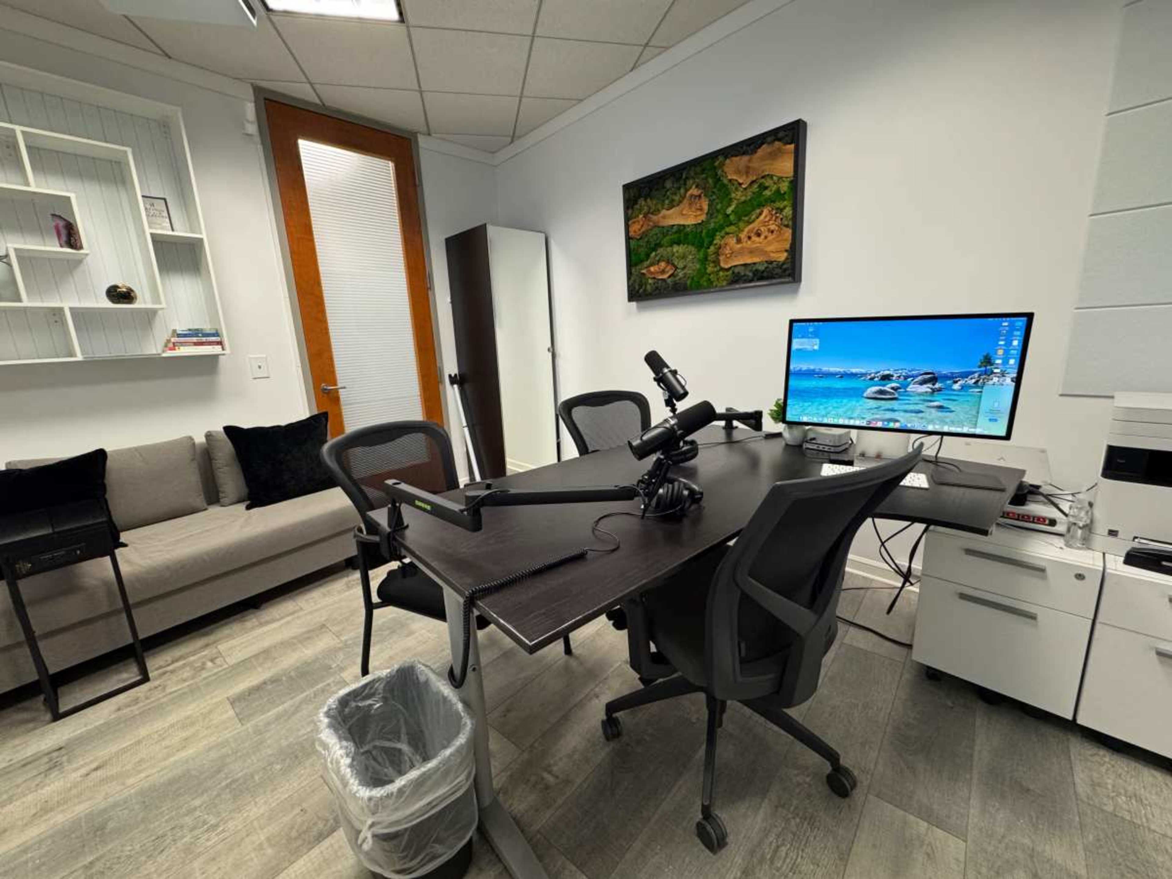The image shows a modern office space featuring a table with four chairs, a computer monitor, a microphone setup, a couch against the wall, and a wastebasket.