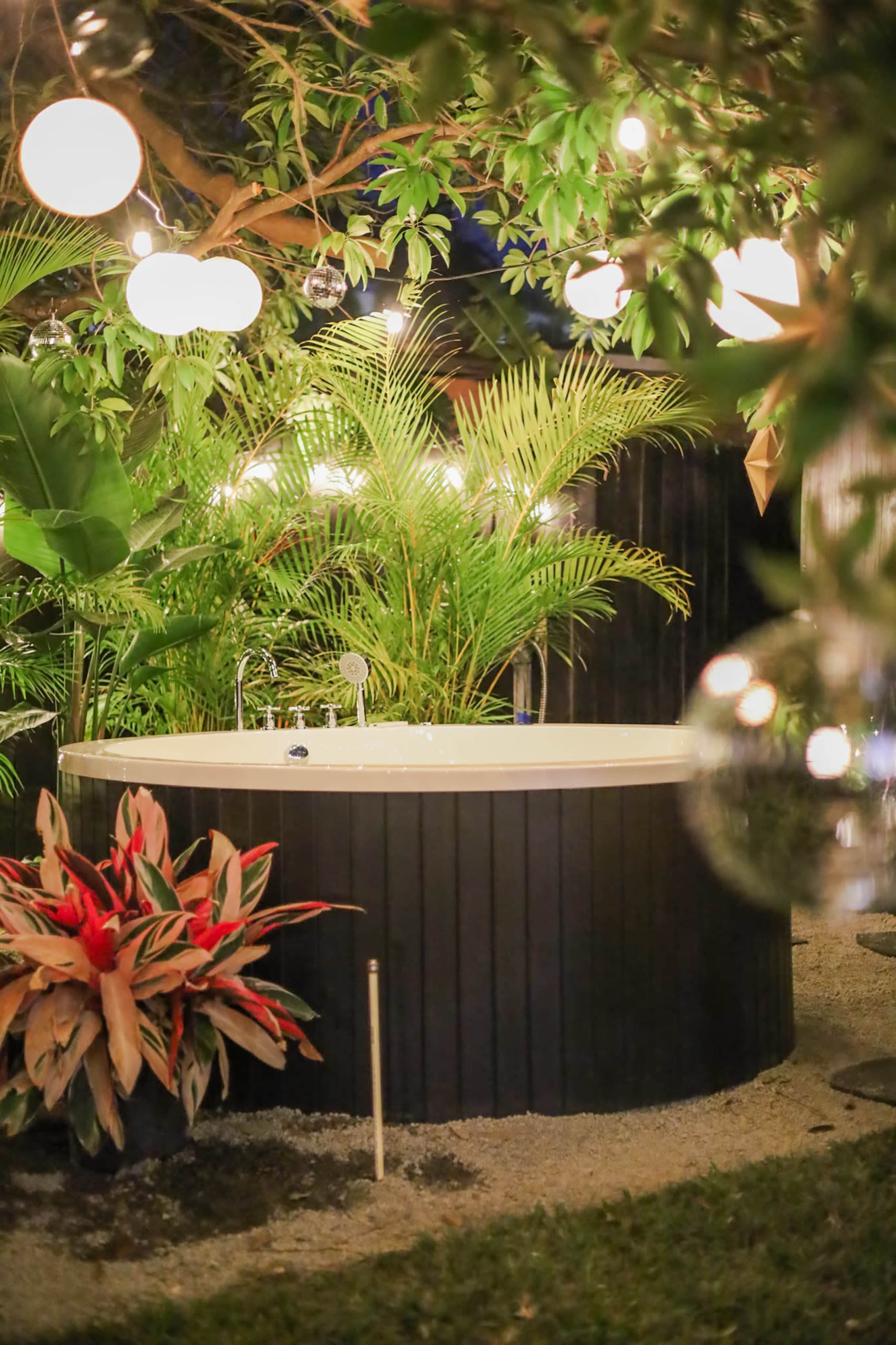 A circular hot tub is surrounded by tropical plants and illuminated by hanging lights in a garden setting.