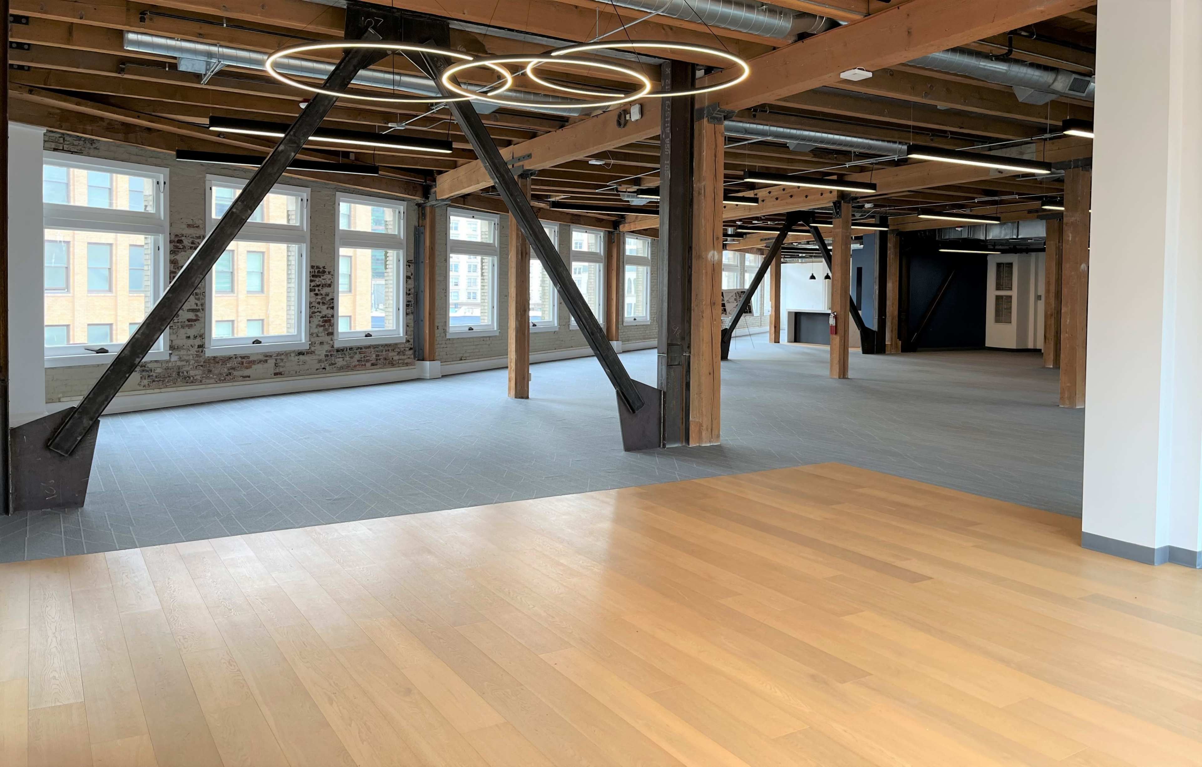 The image shows a spacious, empty room with wooden flooring, exposed beams, and large windows allowing natural light.