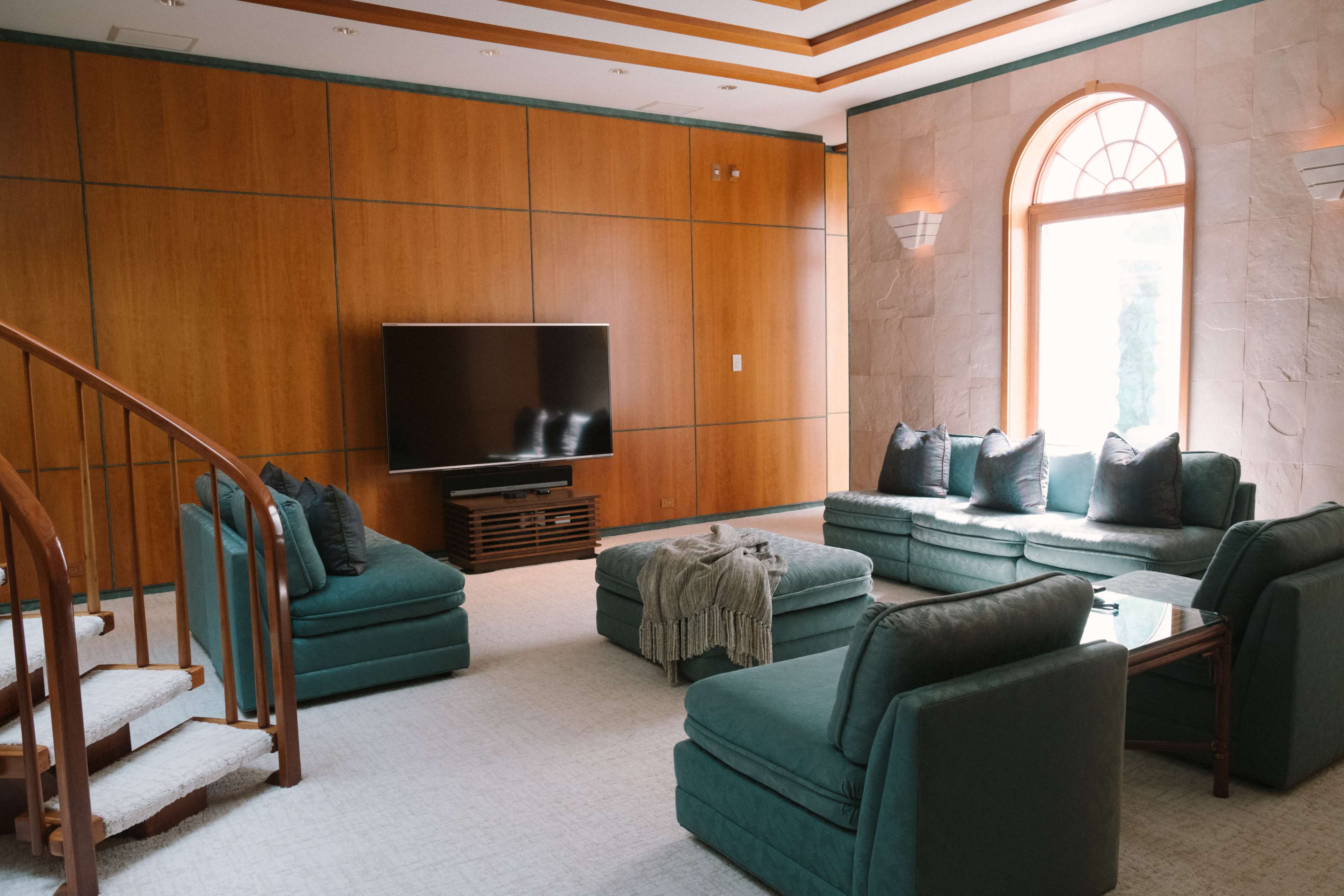 A spacious living room features a large television mounted on a wooden wall, surrounded by teal upholstered furniture and a staircase in the corner.