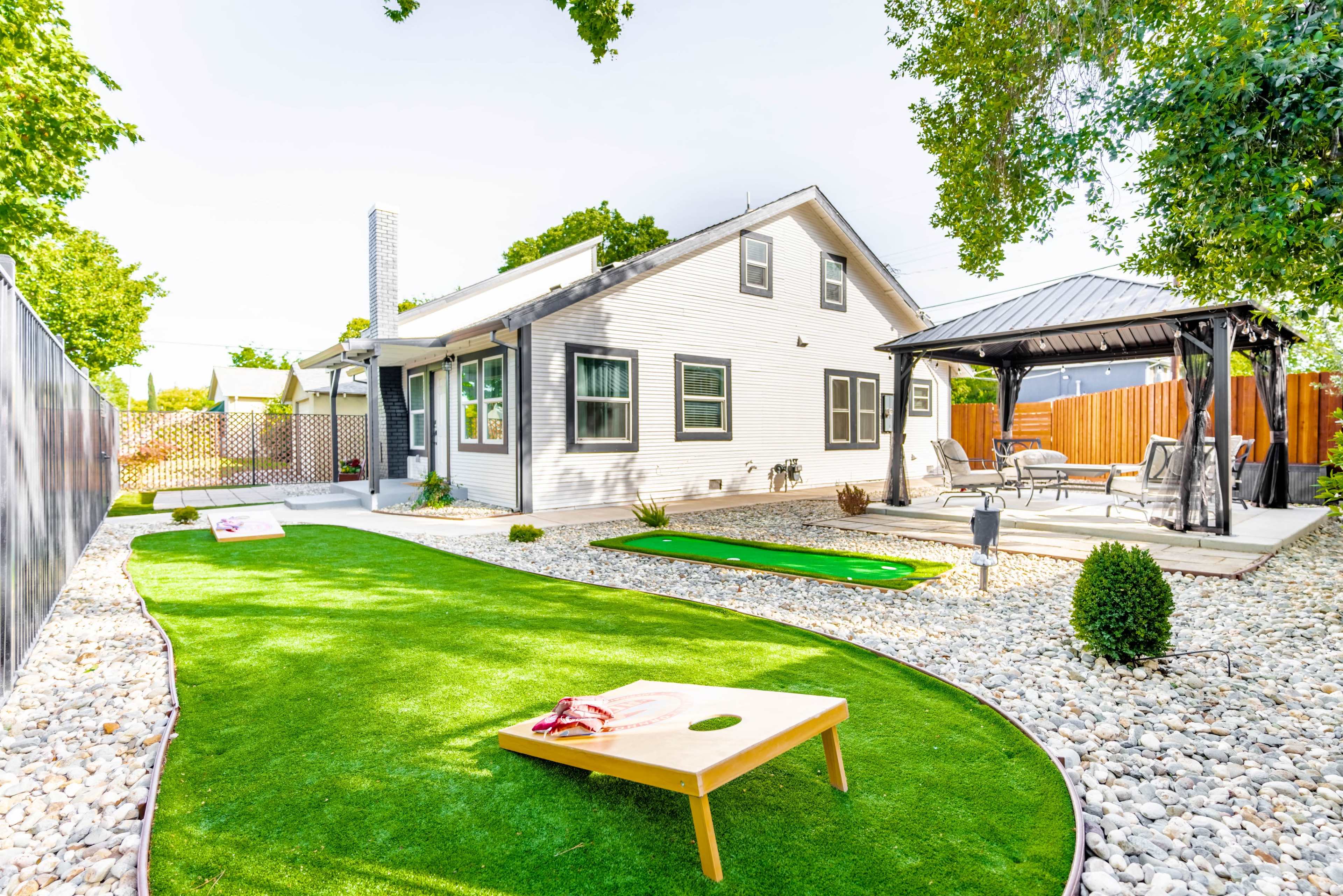 A landscaped backyard featuring a green artificial turf area, a cornhole game set up, a putting green, and a patio with seating.