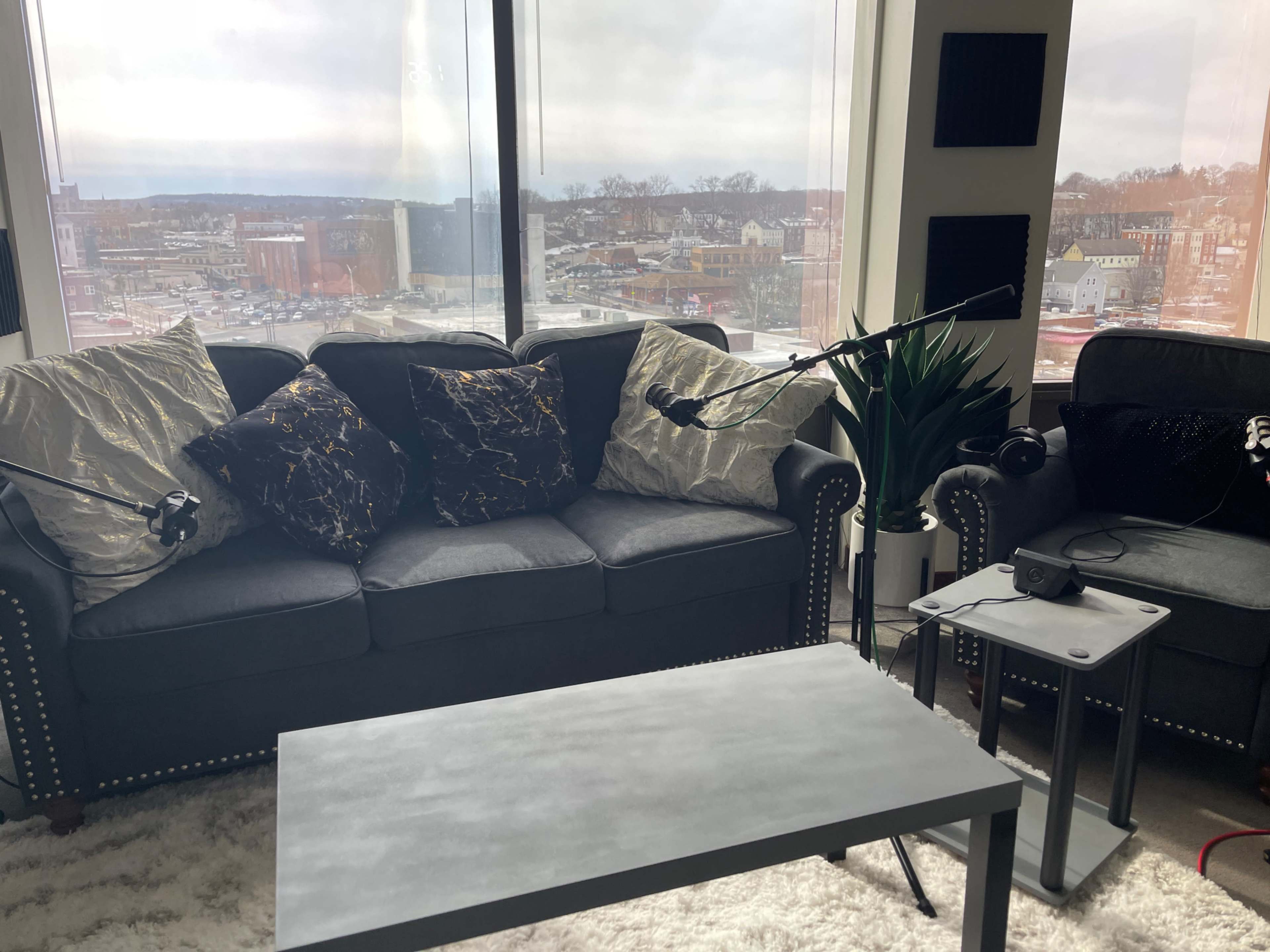 A cozy living room setup with a gray sofa adorned with decorative pillows, a small coffee table, and two microphones positioned for podcasting, all near a large window offering a view of an urban landscape.
