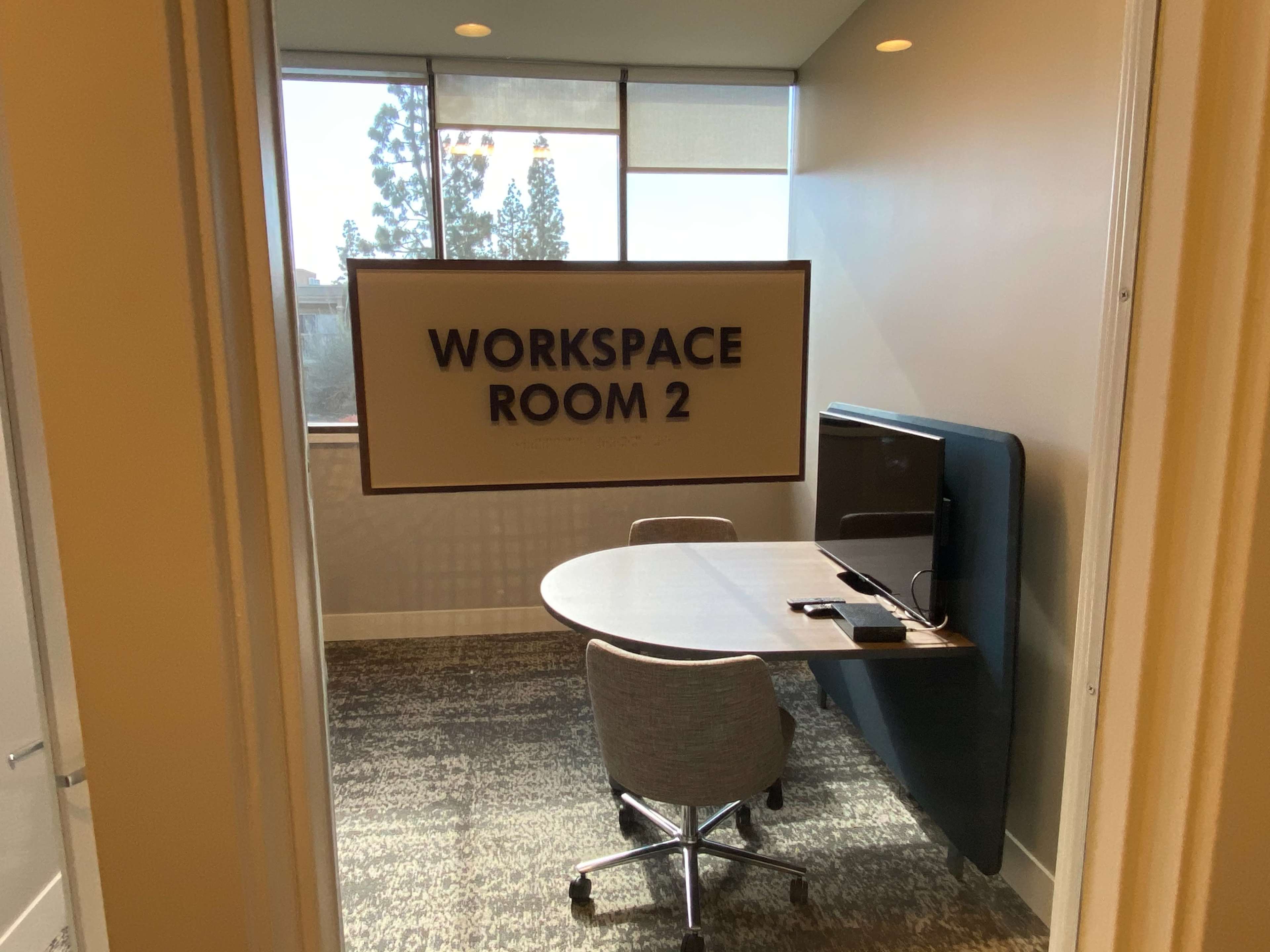 The entrance to a meeting room labeled "WORKSPACE ROOM 2," featuring a round table, two chairs, and a large screen visible inside.