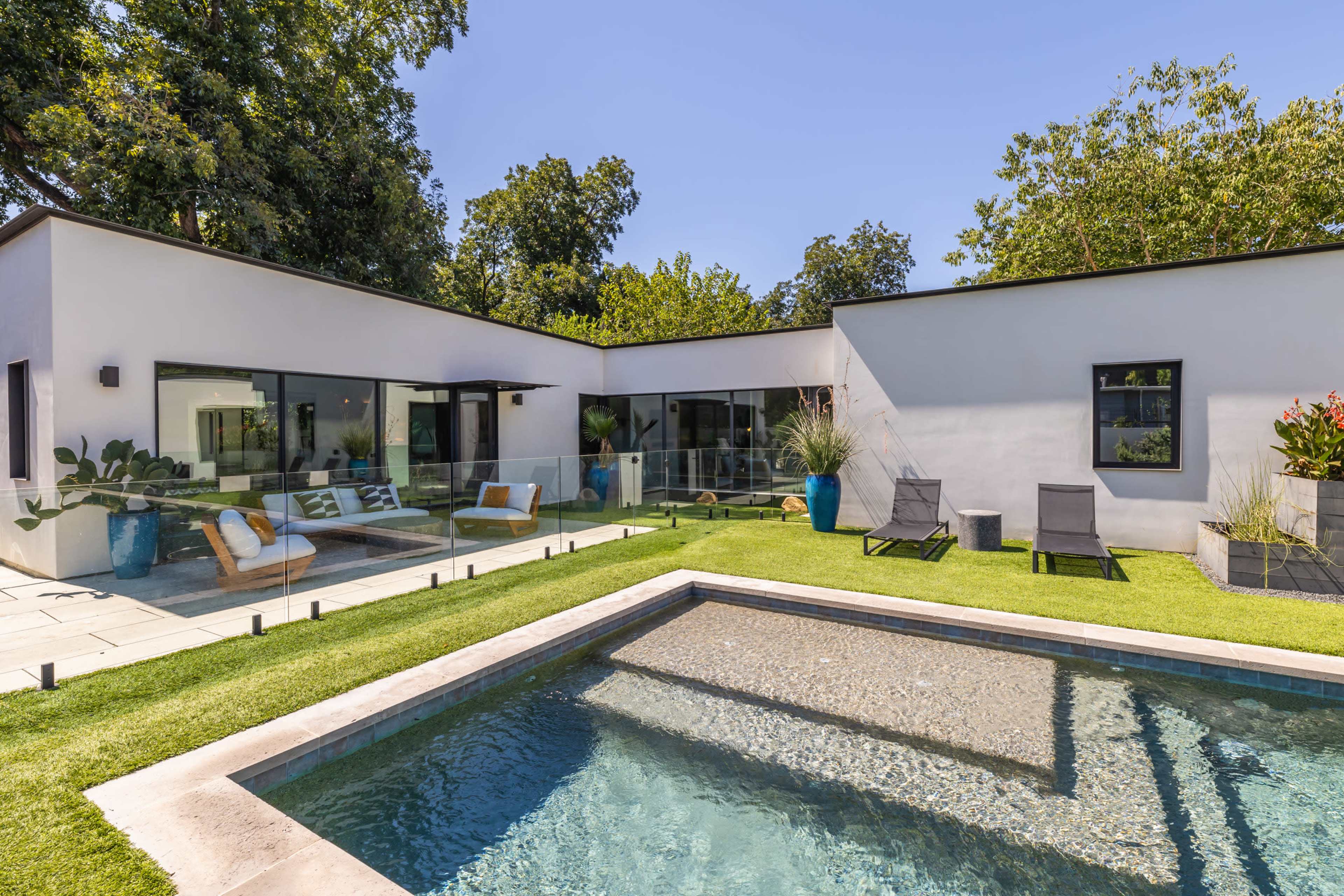 A modern house with large windows surrounding a rectangular swimming pool and a landscaped yard featuring lounge chairs and decorative plants.