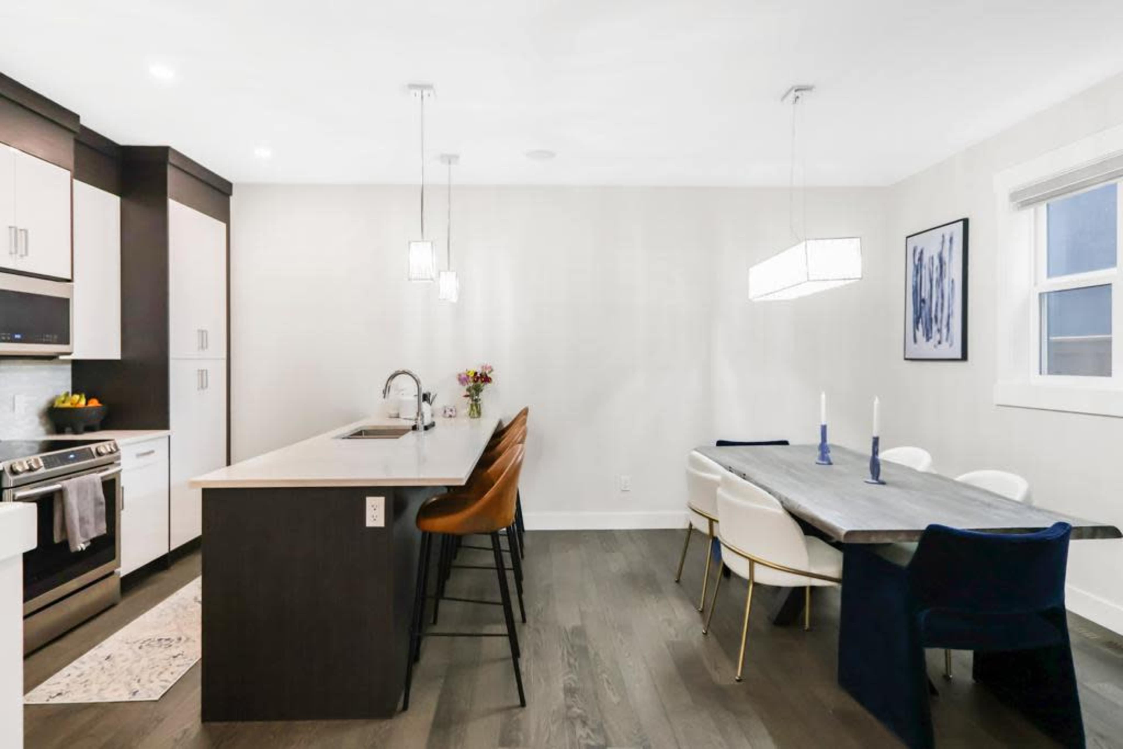 The image shows a modern kitchen and dining area featuring a central island with seating, a formal dining table, and pendant lighting.